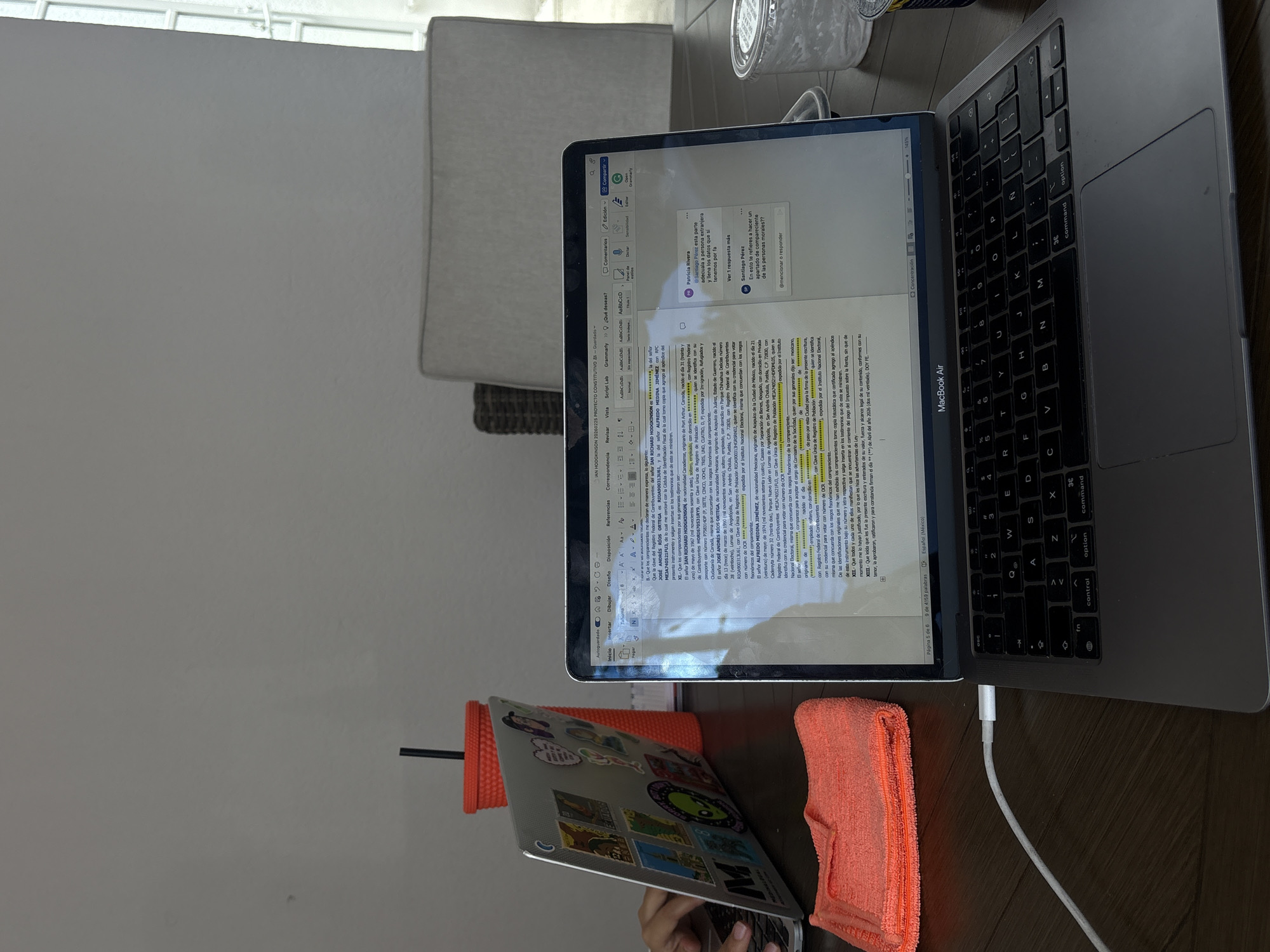 On a wooden surface, a silver MacBook Air laptop is open and displaying a document in Spanish with highlighted text. The laptop is plugged into a white power cable. An orange cleaning cloth rests next to the laptop. A second smaller laptop with stickers on the back props up the larger one. A hand is visible holding the smaller laptop. A window and a light-colored chair are in the background, along with a partially visible plastic container. The document on the screen contains information about people's names, addresses, and RFCs (Registro Federal de Contribuyentes), which suggests a legal or administrative context. The laptop is set to the Spanish (Mexico) language, and the location in San Andrés Cholula, Puebla, Mexico can be identified from the document.