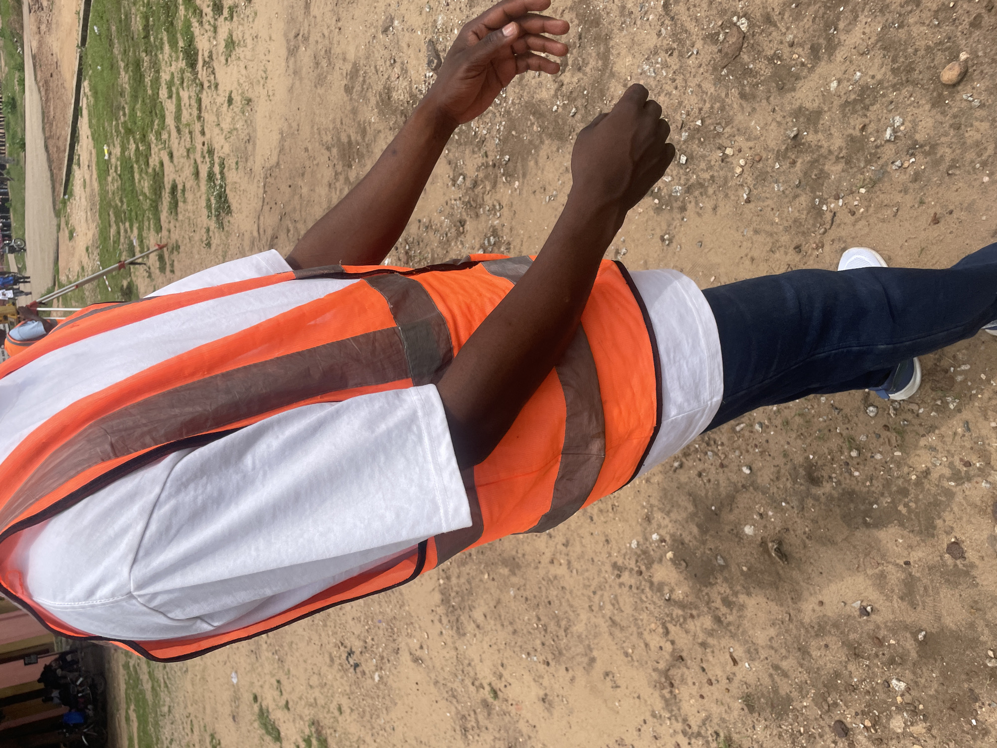 A medium-shot photograph shows the torso and arms of a person wearing a white t-shirt and an orange safety vest with reflective gray stripes. The person is standing on a sandy surface with scattered small stones and patches of dry grass. In the background, a blurry scene depicts other people, some with bicycles and motorcycles, near a building with colorful walls. A surveying tripod is visible in the middle ground. The overall environment suggests an outdoor location in Lokoja, Nigeria, likely a construction site or an open public area.