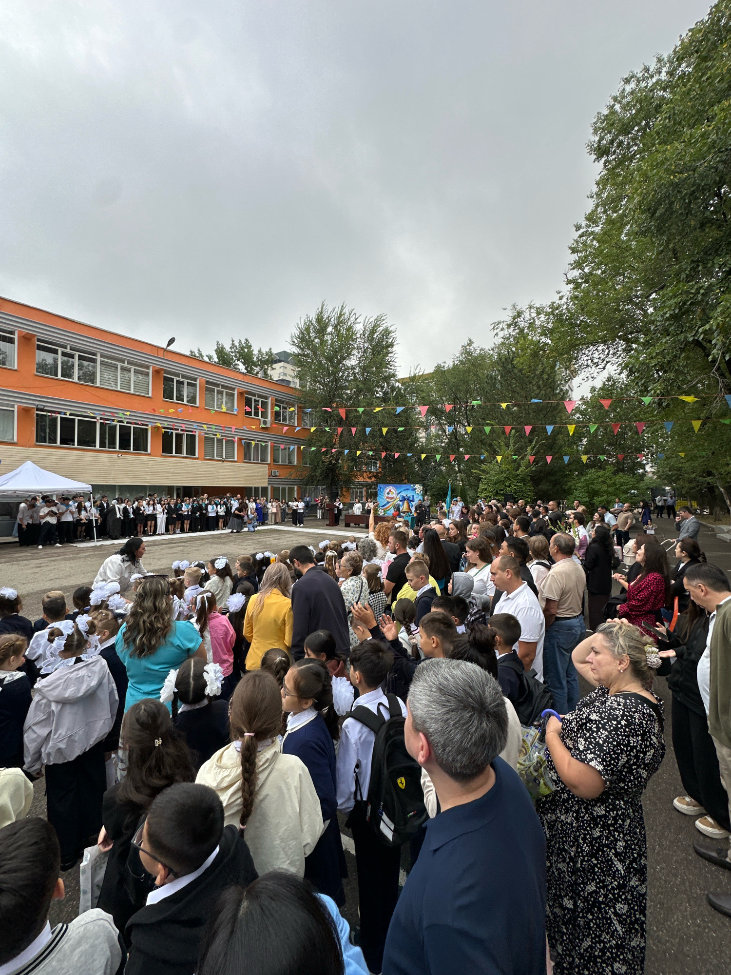 The image depicts an outdoor gathering of people, likely students and adults, in front of an orange-colored school building. The school has multiple stories with rows of windows. Colorful triangular flags are strung across the scene, suggesting a celebratory event. A white tent is set up to the left, and a podium is visible in the distance to the right. The ground appears to be paved, and trees with green foliage are present on the right side of the image. The sky is overcast and gray. People are standing and facing a central area, with some children in the foreground wearing school uniforms and large white bows, a common sight for the first day of school in Kazakhstan. The overall atmosphere suggests a formal school event, possibly the beginning of the academic year, which in Kazakhstan is celebrated on September 1st. The architecture of the building and the presence of Kazakh flags in the distance hint at the location being Almaty, Kazakhstan.