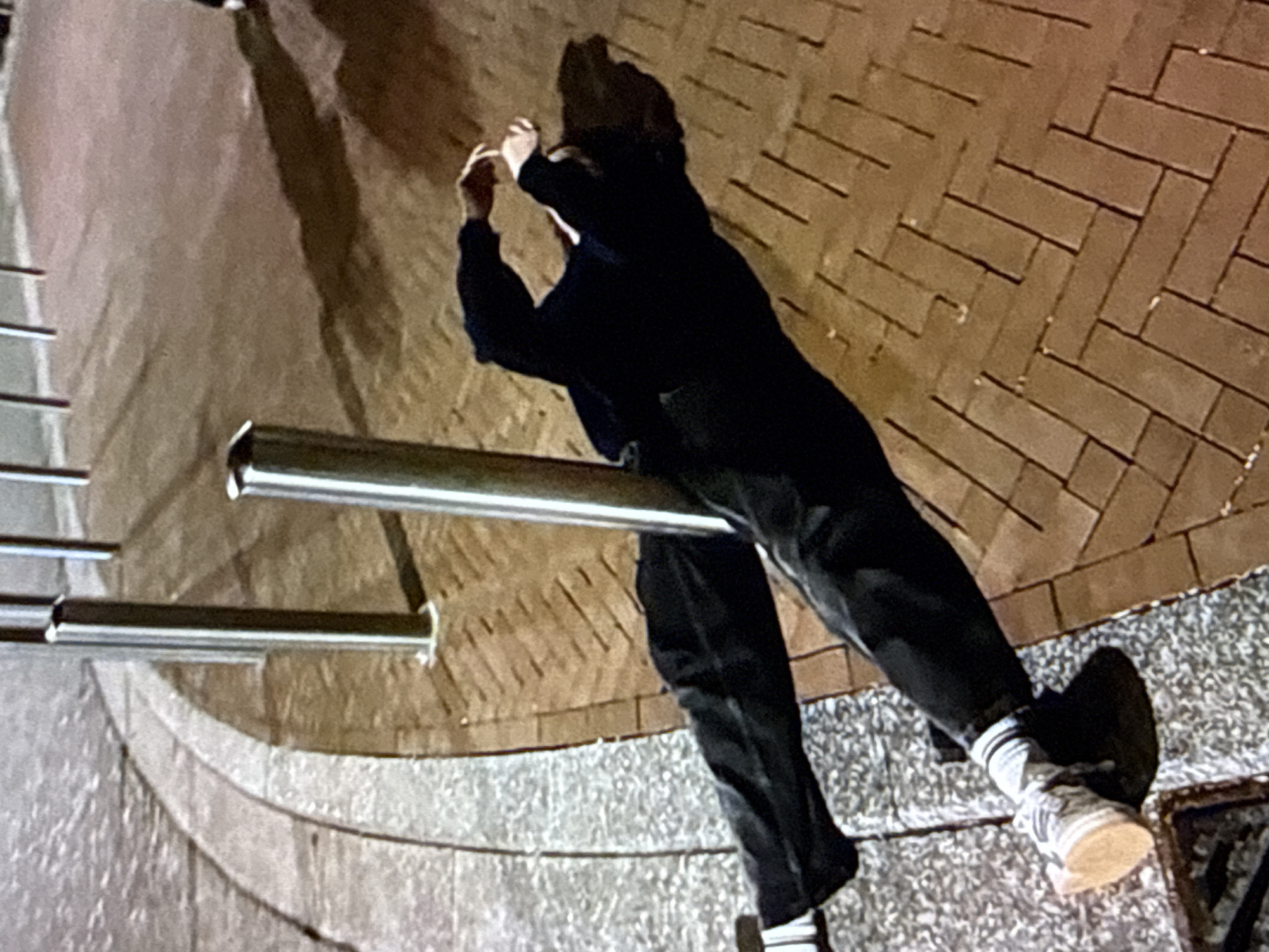 An individual is depicted lying on the ground in an outdoor urban environment, likely under artificial lighting conditions at night. The person is positioned horizontally, with their back against a silver-colored metal bollard. Their feet rest on a raised concrete curb, separating a textured light-grey aggregate surface (possibly a road or another sidewalk section) from a reddish-brown brick paver sidewalk laid in a herringbone pattern. The individual is wearing dark trousers, white socks with dark stripes, and light-colored sneakers with prominent soles. They are also wearing a dark long-sleeved top or jacket. Their head is turned away from the camera, and their arms are bent with hands near their head. A distinct shadow of the individual and the bollards is cast on the brick pavement, indicating a light source from the upper left of the frame. Multiple other similar bollards are visible in a linear arrangement in the background. No identifying markers confirming the specific location of Kardzhali, Bulgaria are visible in the image.