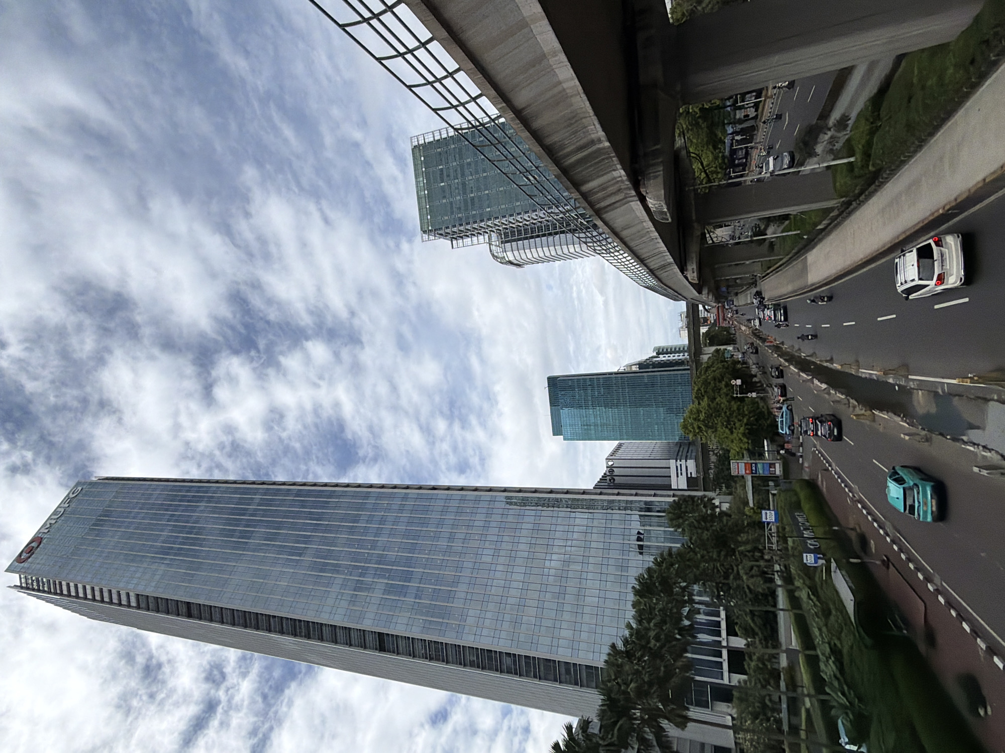 The image shows a view from a low angle, looking up at skyscrapers and an elevated highway in South Jakarta, Indonesia. The sky is partially cloudy. A tall, rectangular skyscraper with a glass facade dominates the left side of the frame. Another glass-clad skyscraper is partially visible. Palm trees line the road below. A highway runs along the right side of the image with vehicles visible. An elevated rail line with a trellis-like structure passes overhead.