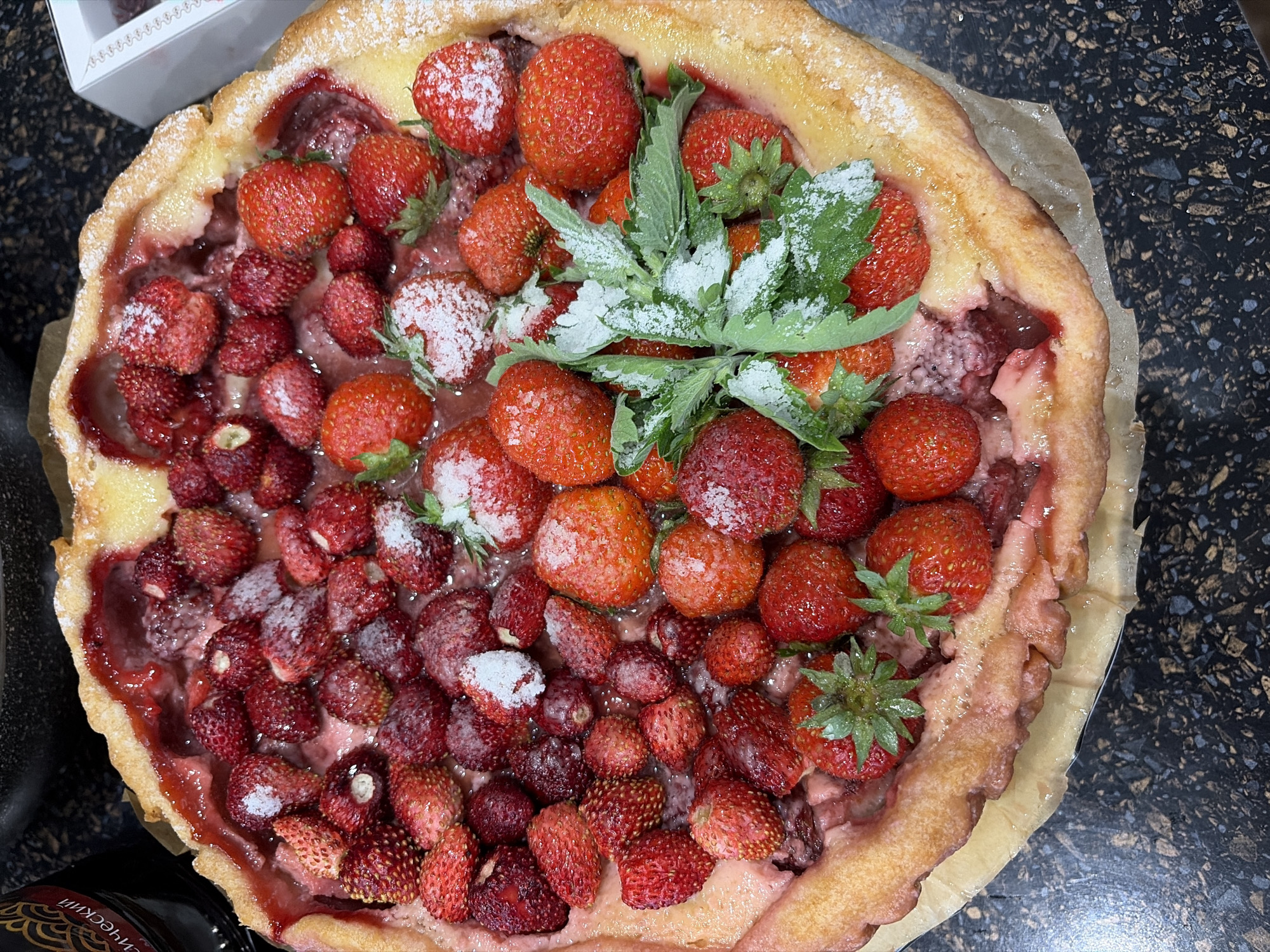 A high-angle, close-up shot captures a rustic strawberry tart, likely homemade, presented on a dark, speckled countertop. The tart is generously filled with a mix of ripe strawberries, ranging in size from small wild strawberries to larger cultivated ones. Some of the berries are dusted with powdered sugar, giving them a frosted appearance. A few sprigs of fresh mint, also lightly coated in sugar, are placed in the center as a garnish. The crust of the tart is golden brown and appears slightly uneven, suggesting a homemade quality. The filling is a vibrant red, indicating the presence of strawberry jam or cooked strawberries.
In the upper left corner, a partially visible white box with a decorative pattern is present, hinting at a possible purchase or presentation of baked goods. The overall scene is indoor, with the dark countertop providing a strong contrast to the bright colors of the fruit and crust. The lighting is natural, creating soft shadows and highlighting the textures of the tart. There are no people visible in the frame. The image is devoid of any specific time of day or weather cues. No other discernible location cues are present beyond the countertop. The image evokes a sense of homemade comfort and the freshness of summer fruits. There is partially visible text on a dark label in the bottom left corner, but it is too indistinct to be read. The provided location context of "Северка, Russia" does not offer specific visual information within the image.