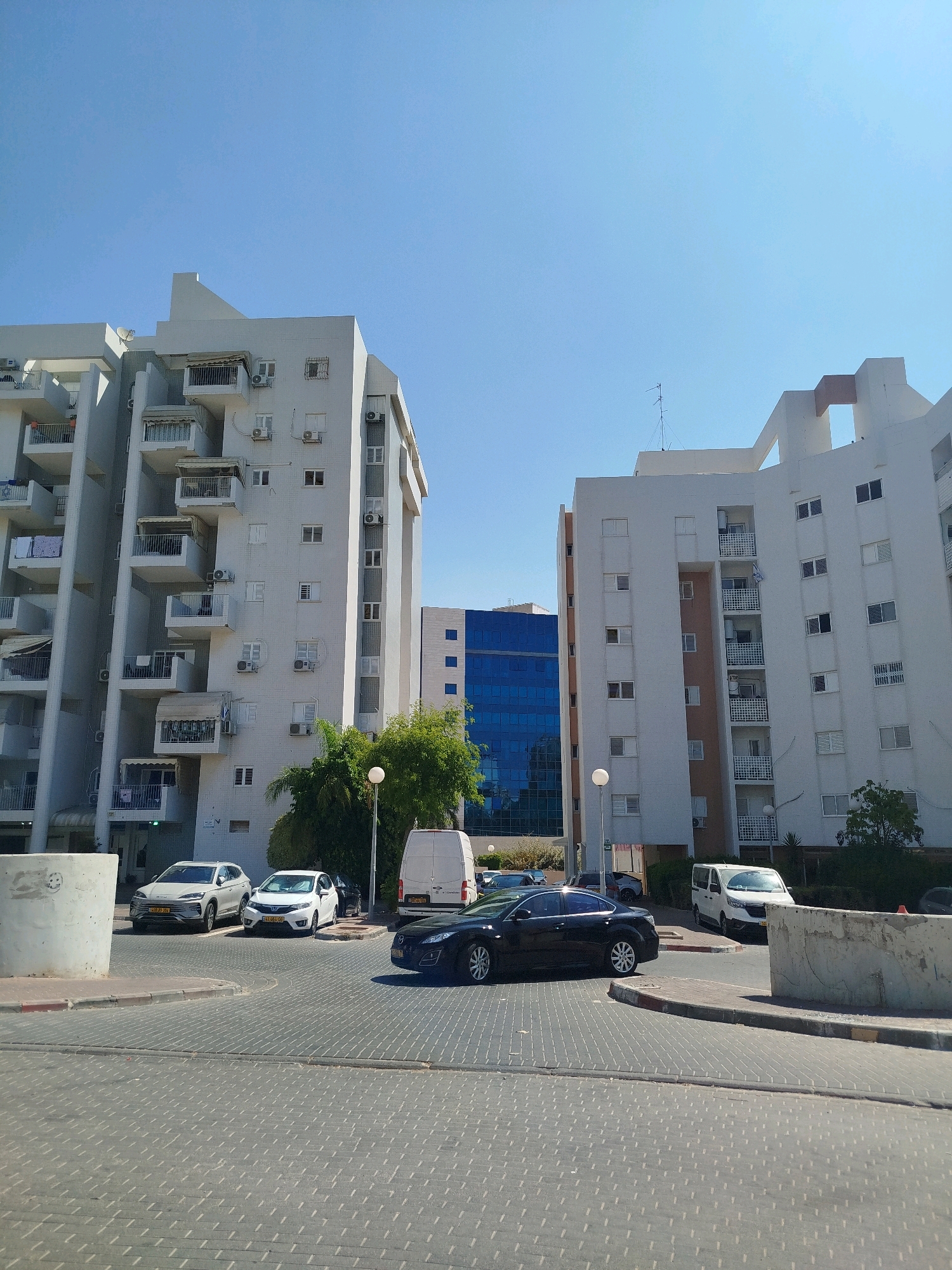 A street view in Ashkelon, Israel, captures a modern urban scene under a clear blue sky. In the foreground, a paved road with a distinctive interlocking brick pattern leads towards two large residential buildings. The building on the left is a multi-story white structure with numerous balconies, some of which have clothes drying on them. The building on the right is similar in design, also white, with multiple floors and balconies. Between these buildings, a modern blue glass office building is visible. Several cars are parked or driving on the street, including a white minivan, a white sedan, a grey SUV, and a black sedan. A street lamp stands near the white minivan. The environment appears to be well-maintained and residential.