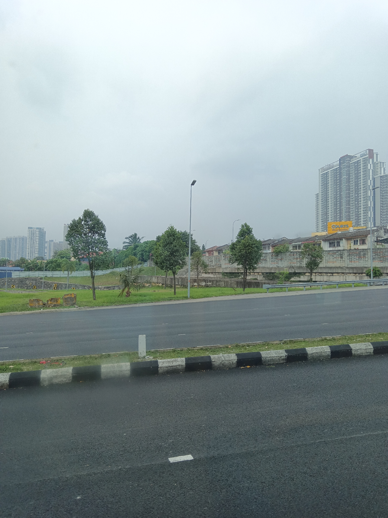 The image depicts a roadside view in a city, likely Kuala Lumpur, Malaysia, under an overcast sky. In the foreground, a dark asphalt road with white lane markings is visible, bordered by a curb with alternating black and white concrete blocks. Beyond the curb, a grassy median separates the road from a concrete wall with a metal guardrail.
A line of houses with tiled roofs is situated behind the concrete wall. Further in the background, several trees are interspersed with the houses and a large, multi-story building with a sign that reads "COURTS" in yellow. To the left and right of this building, numerous high-rise apartment buildings dominate the skyline, some appearing to be under construction. The overall impression is one of urban development with a mix of residential and commercial structures. The weather appears to be humid and possibly rainy, given the wetness of the road surface. No people or vehicles are clearly visible in the frame.