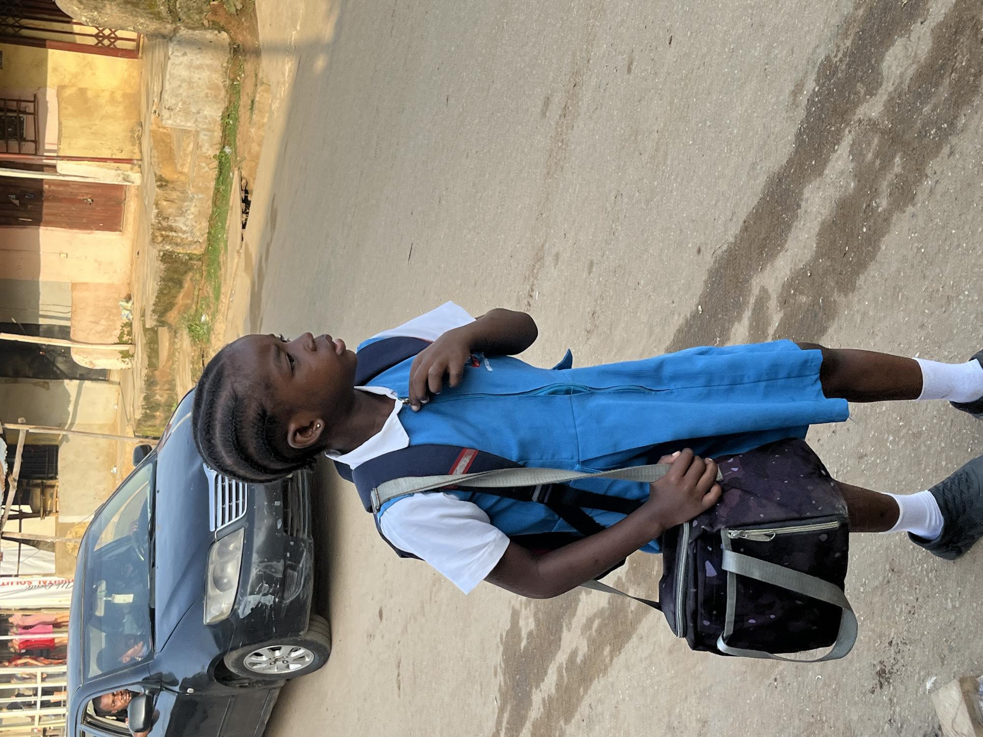 A young girl wearing a blue school uniform with a white shirt and white socks stands on a paved street. She has braided hair and is carrying a dark purple lunch bag with a gray strap over her left shoulder. She is looking to her left, with her right hand holding onto the strap of her lunch bag. A black car is parked to her left. The street appears to be in a somewhat undeveloped urban area, with buildings made of concrete and corrugated metal visible in the background. Sunlight suggests it is daytime. The image's orientation is rotated 90 degrees clockwise.