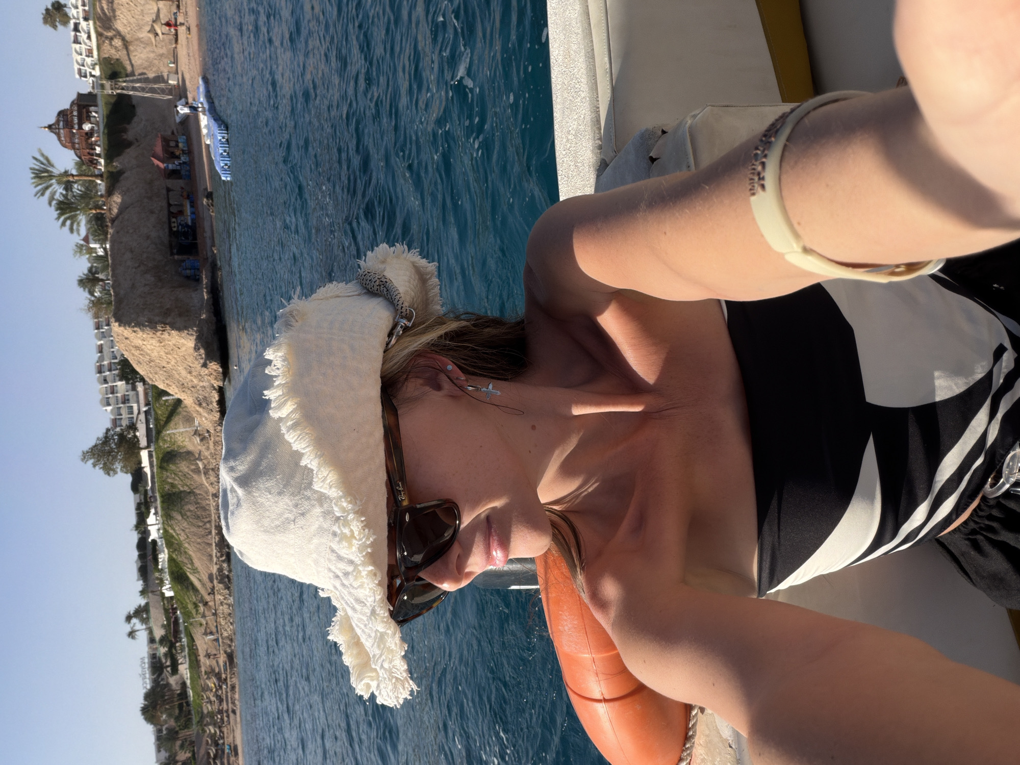 A woman captures a selfie while enjoying a sunny day on a boat. She is depicted from the chest up, wearing dark sunglasses with the "Ray-Ban" logo visible on the arm, a light-colored fringed sun hat, and a black and white patterned top, possibly a swimsuit. A delicate cross-shaped earring and a light-colored bracelet are also noticeable. Her hair is long and brown, and her skin appears tanned.
She is situated on a boat, with a portion of an orange lifebuoy visible beside her. The boat floats on clear, blue water, likely the sea, which shows ripples and reflections of the bright sunlight.
In the background, a picturesque coastline unfolds under a clear sky, suggesting a resort environment. The sandy shoreline is adorned with green palm trees and several multi-story buildings, characteristic of hotels or resorts. One prominent building with a domed roof stands out on the left. Beach amenities, possibly sun loungers or umbrellas, are visible along the shore. A sign in the distance, though partially obscured, appears to read "MOVENPICK". The setting sun casts a warm glow, indicating it's either late morning or afternoon. The overall scene conveys a relaxed, vacation atmosphere in a warm, sunny location, consistent with Sharm El Sheikh, Egypt.