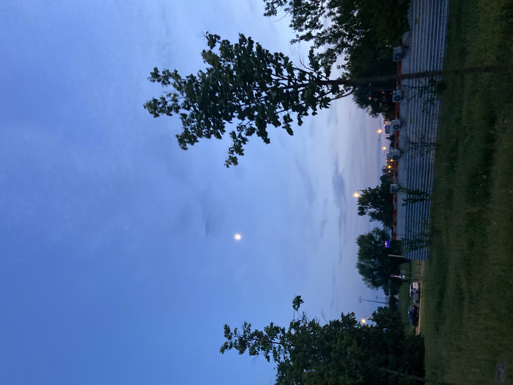 The image captures a dusky sky over a suburban or semi-urban landscape. The sky is a gradient of deep blue at the top fading to lighter hues near the horizon, with scattered wispy clouds. A full moon is visible in the upper right quadrant of the sky. In the foreground, there is overgrown grass. Several trees of varying heights and densities are present, with a prominent tree in the middle right of the frame. In the middle ground, a construction barrier made of corrugated metal siding runs horizontally across the image, topped with what appear to be multiple air conditioning units. Beyond the barrier, several buildings with lit windows and streetlights are visible, indicating a developed area. Two cars, a dark SUV and a lighter colored SUV, are parked on a paved surface to the left, suggesting the presence of a road or parking lot. The overall impression is a quiet evening scene, possibly on the outskirts of a city. Given the visual cues of residential buildings, construction, and a generally maintained landscape, this could be a scene from a less dense area within Warsaw, Poland, possibly a developing district or a park bordering a residential zone.