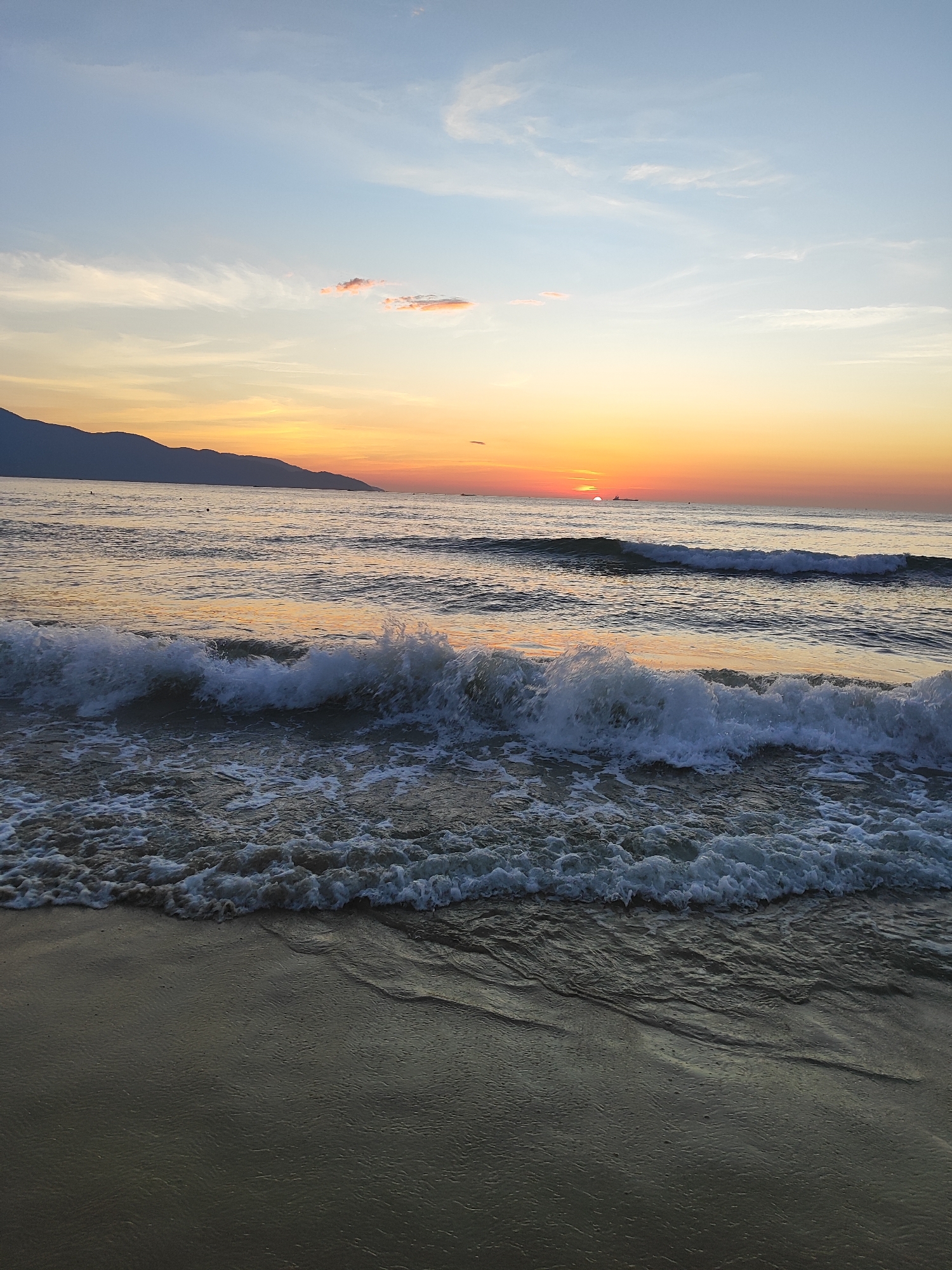 The image depicts a serene sunrise over the ocean at My Khe Beach in Da Nang, Vietnam. The sky transitions from a soft blue at the top to warm hues of orange and yellow near the horizon, where the sun is just beginning to rise. Wispy clouds are scattered across the sky, some tinged with pink by the sunrise. In the distance, a silhouetted mountain range forms the coastline. The ocean is calm, with gentle waves rolling towards the shore, creating white foam as they break. The wet sand in the foreground reflects the colors of the sky, and a small portion of the beach is visible at the bottom of the frame. There are no people visible in the image, only a few distant boats or ships on the water. The overall atmosphere is peaceful and tranquil, characteristic of an early morning at this popular Da Nang beach.