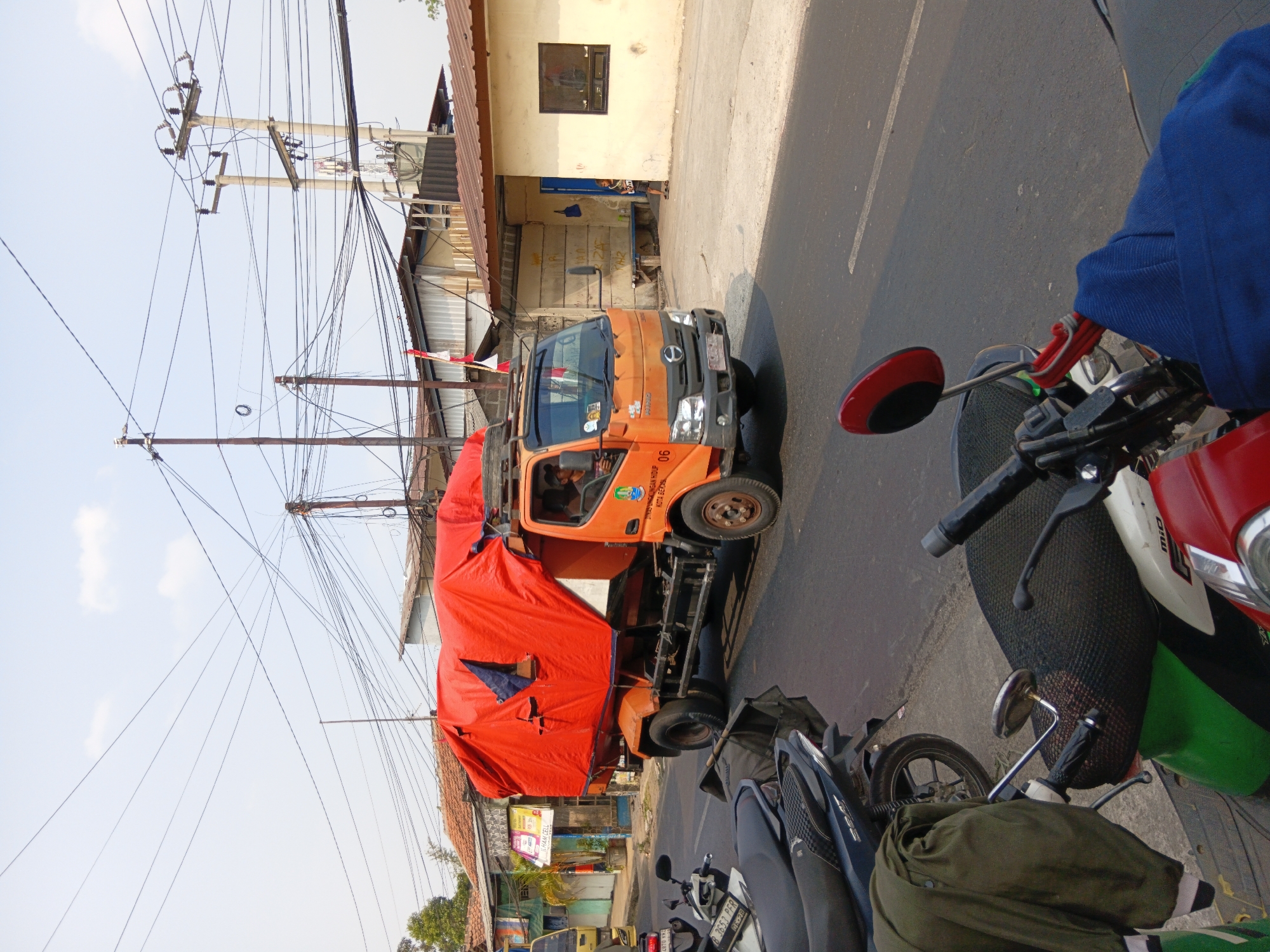 An orange dump truck, covered with a bright orange tarp and a dark blue tarp, is driving on a street in Bekasi, Indonesia. The truck has a license plate that reads "B 9056 KCA". A yellow truck is parked to the left of the dump truck. Several motorcycles are parked on the side of the road. In the background, there are buildings with tiled roofs, power lines, and a few trees. The sky is clear and blue. The image appears to have been taken on a sunny day.
