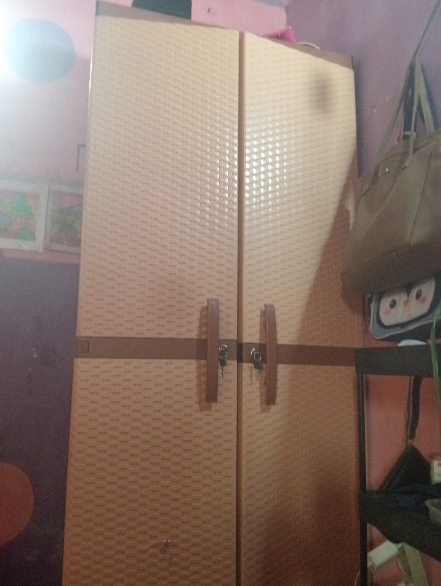 This is a medium-shot, indoor photograph of a beige plastic wardrobe with two doors. The doors have a textured, woven pattern and are secured by two dark brown handles, each with a small silver lock. The wardrobe is positioned against a pink wall. To the right of the wardrobe, a dark brown shelf unit holds a light brown handbag hanging from the top, and a grey bag with an owl design. The environment suggests a bedroom or living space. The image appears to be taken in Bloro, Indonesia, based on the context provided.