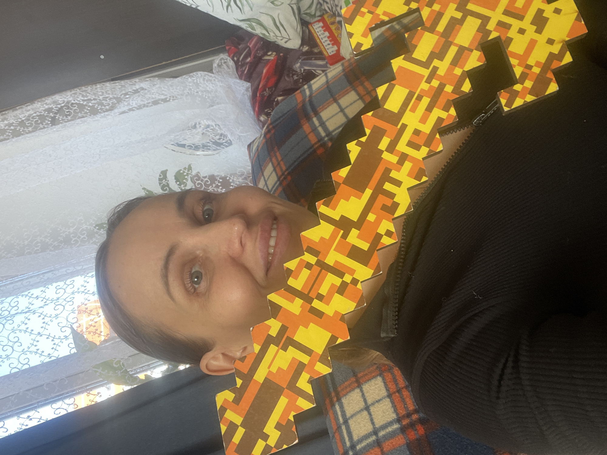 A woman is photographed indoors. She has fair skin, brown hair pulled back, and is smiling slightly. She is wearing a black ribbed shirt or jacket. A zig-zag shaped object, patterned with yellow, orange, and brown geometric shapes, is positioned in front of her, partially obscuring her face. A plaid blanket or scarf in shades of blue, brown, and orange is draped across her chest and shoulder. In the background, there is a lace curtain and patterned bedding with a pillow featuring green leaf designs. A package of some sort is also visible in the background. I cannot verify the location is Chaikovsky, Russia.