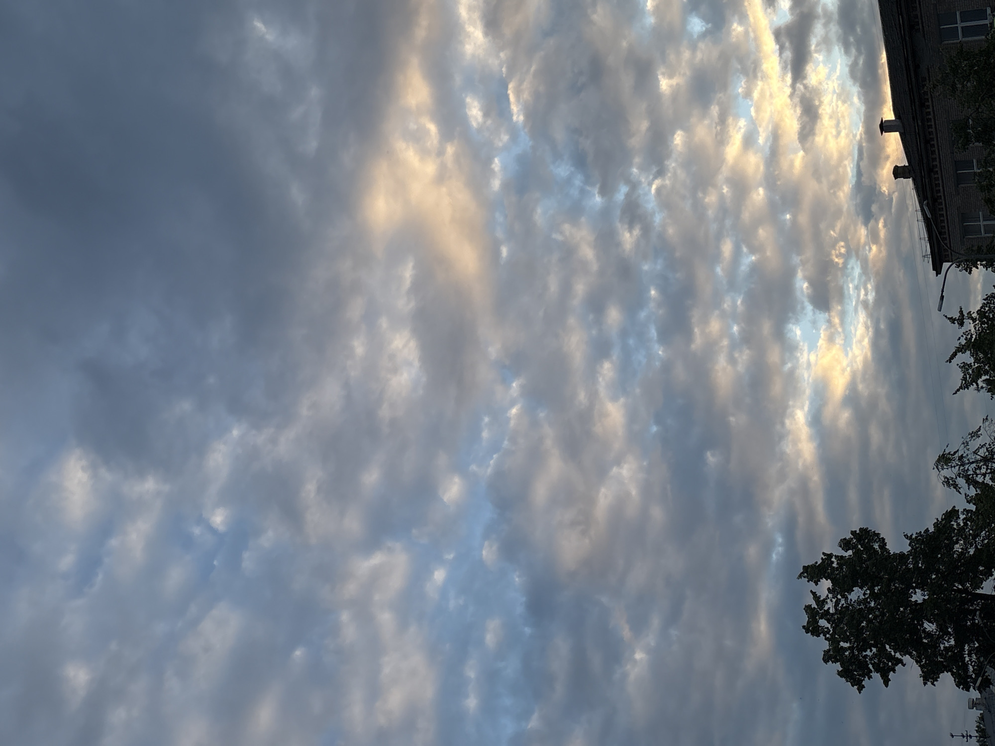 This is a photograph of a cloudy sky above an urban environment in Chaikovsky, Russia. The sky is filled with a mix of gray and white clouds, with some patches of light blue sky visible. Sunlight is breaking through the clouds in some areas, casting a warm, golden glow. In the foreground, the tops of trees and the roof of a building are visible. The building appears to be made of brick and has a dark roof with chimneys. There are no people or animals visible in the frame. The overall impression is of a dynamic and atmospheric sky, possibly at dusk or dawn.