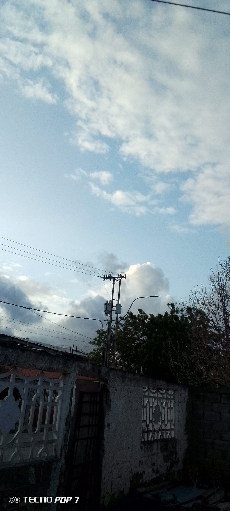 The image depicts an outdoor scene in Altagracia, Venezuela. The sky is mostly clear with scattered white clouds. In the mid-ground, there's an electrical pole with power lines running across the frame. Below the power lines, there's a low, weathered structure with a section of decorative concrete lattice, a dark doorway, and a window with a metal grate. To the right of the structure, there are trees with bare branches and dense green foliage. The light suggests it's daytime. A "TECNO POP 7" watermark is visible at the bottom left corner.