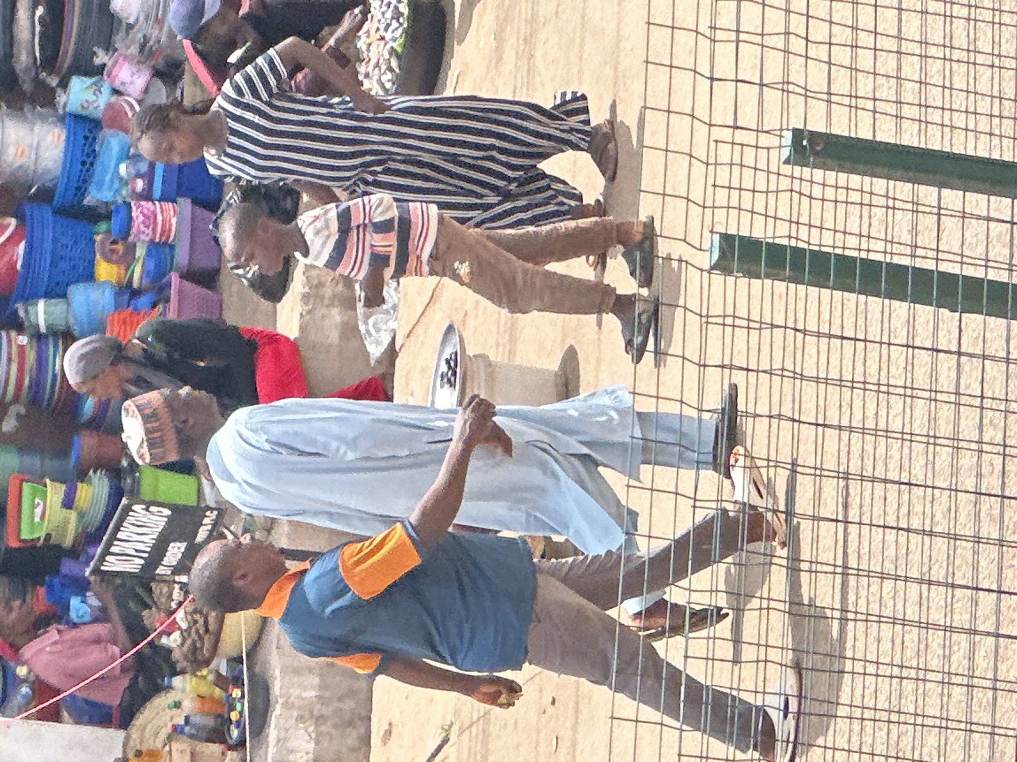 The image depicts a street scene in Bukuru, Nigeria, characterized by a bustling marketplace atmosphere. A group of people are walking on a dusty ground, possibly a market street.
In the foreground, a man wearing a blue and orange t-shirt and brown trousers is walking from left to right. He is followed by another man dressed in a light blue traditional garment, also walking from left to right. To the right of these two men, a young boy in a striped shirt and brown trousers walks in the same direction. A woman in a black and white striped dress is also visible on the right side of the frame, walking towards the right.
Behind the people, various market stalls are set up, displaying a variety of goods. These include colorful plastic buckets and containers of different sizes, as well as some food items, possibly fruits or vegetables, in baskets. A "NO PARKING BY ORDER" sign is prominently displayed on a black board, indicating regulations within the area. The overall environment suggests a typical Nigerian market setting with a lively and active scene. The image is captured from a slightly elevated angle, looking down on the subjects and the market. The presence of a wire fence in the foreground suggests a designated viewing area or a barrier.