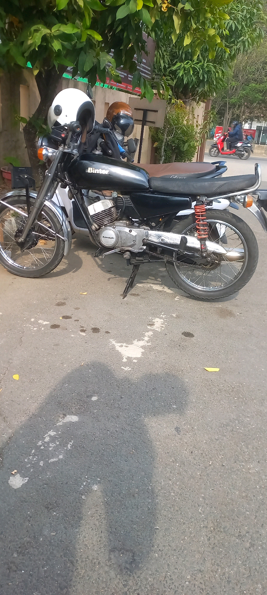 A black Binter motorcycle is parked on a paved road in Samarinda, Indonesia. Two helmets, one white and one brown, are placed on the handlebars of the motorcycle. The motorcycle has a visible Kawasaki engine. In the background, a red scooter is seen driving on the road. Trees and foliage are visible behind the motorcycle, and a building with signs can be seen in the distance. The road surface appears to be asphalt with some white patches and oil stains. A shadow of the motorcycle and possibly a person is cast on the road.