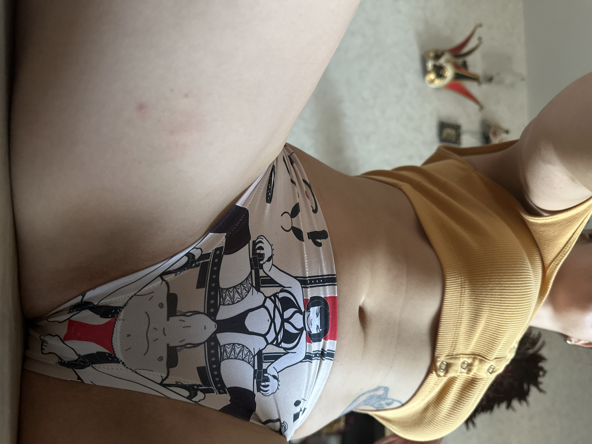 The image shows a close-up of a person's midsection, wearing a yellow ribbed crop top and patterned underwear. The underwear features a graphic of a woman in a chair and another figure below. In the blurred background, a decorative mask and other objects are visible on a wall. The image is taken from a low angle, looking upwards towards the person. No specific location in Vladimir, Russia, is identifiable from the image.