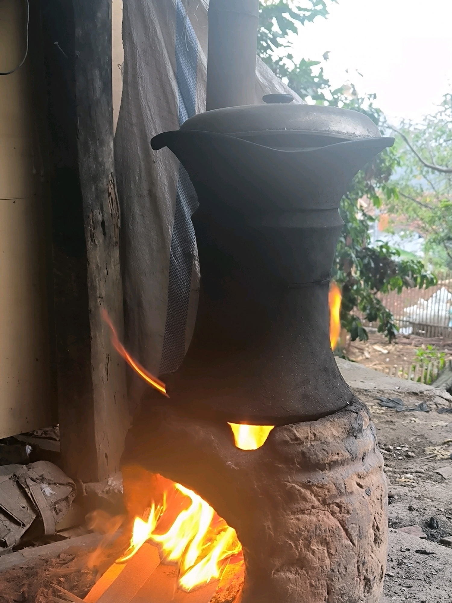 In the village of Cilengkrang, Indonesia, an outdoor cooking scene unfolds. A traditional clay stove, rough and textured, supports a large, dark pot with a lid. Flames erupt from the open base of the stove, casting a warm, orange glow on the surrounding area. To the left, a weathered wooden post stands beside a draped white and blue cloth. Beyond the stove, lush green foliage and a fence made of horizontal wooden planks suggest a rural or semi-rural setting. The ground is covered in dirt and debris. The image captures a moment of traditional cooking in a rustic outdoor environment.