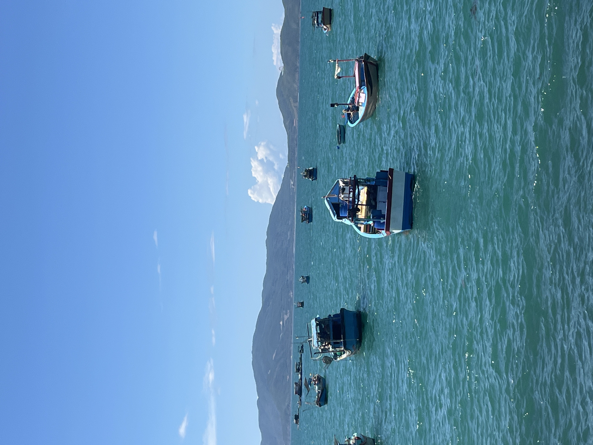 The image shows several fishing boats in the sea, likely located near Bac Nha Trang Ward, Vietnam, based on the environment. The boats are various sizes and colors, appearing to be anchored or drifting gently. A coastline with mountains can be seen in the distance. The sky is blue with some clouds. The water is a turquoise color, suggesting a clear, shallow sea.