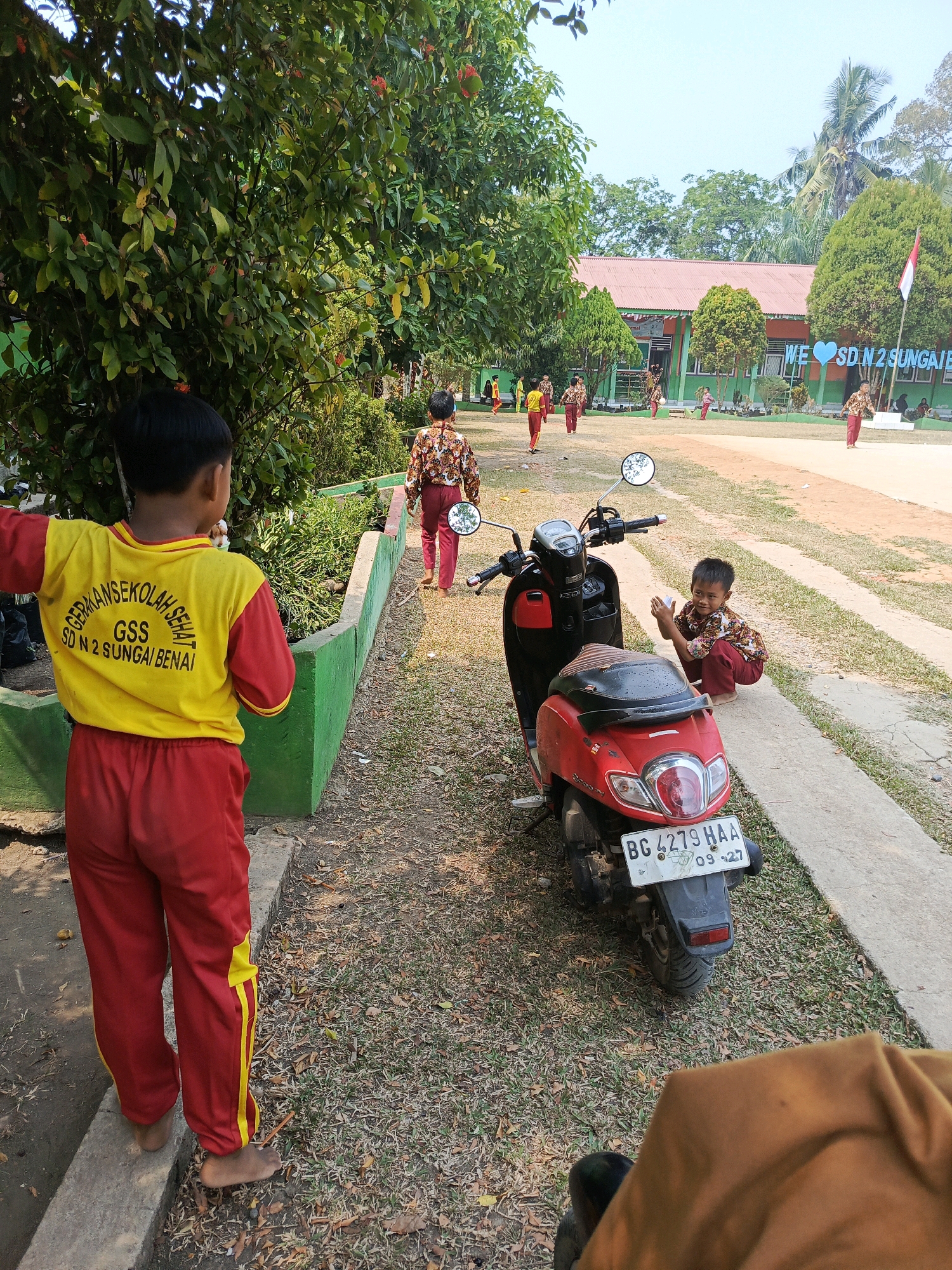 In the city of Megang Sakti IV, Indonesia, a group of young students is captured in a moment of leisure on the grounds of SD N 2 SUNGAI BENAI. The scene unfolds under a bright, clear sky, with lush green trees and tropical foliage framing the school building. A red and black scooter is parked on a concrete path, its license plate reading "BG 4279 HAA."
In the foreground, a boy wearing a yellow and red school uniform, with the words "GERAKAN SEKOLAH SEHAT GSS SD N 2 SUNGAI BENAI" printed on his shirt, stands facing away from the camera. He is barefoot and appears to be observing the activity around him. To his right, another boy, dressed in a patterned shirt and red pants, is crouching near the scooter, his hands clasped in front of him as if in mid-conversation or playful engagement.
Further in the background, several other students in similar school uniforms are scattered across the grassy field and the paved area in front of the school building. Some are walking, while others appear to be interacting with each other. The school building itself is a single-story structure with a red roof and green walls, adorned with colorful banners and an Indonesian flag flying from a flagpole. The overall atmosphere suggests a lively school environment during a break or informal activity. The ground is a mix of dry grass, dirt, and concrete pathways, indicating an outdoor setting typical of tropical Indonesian schools.