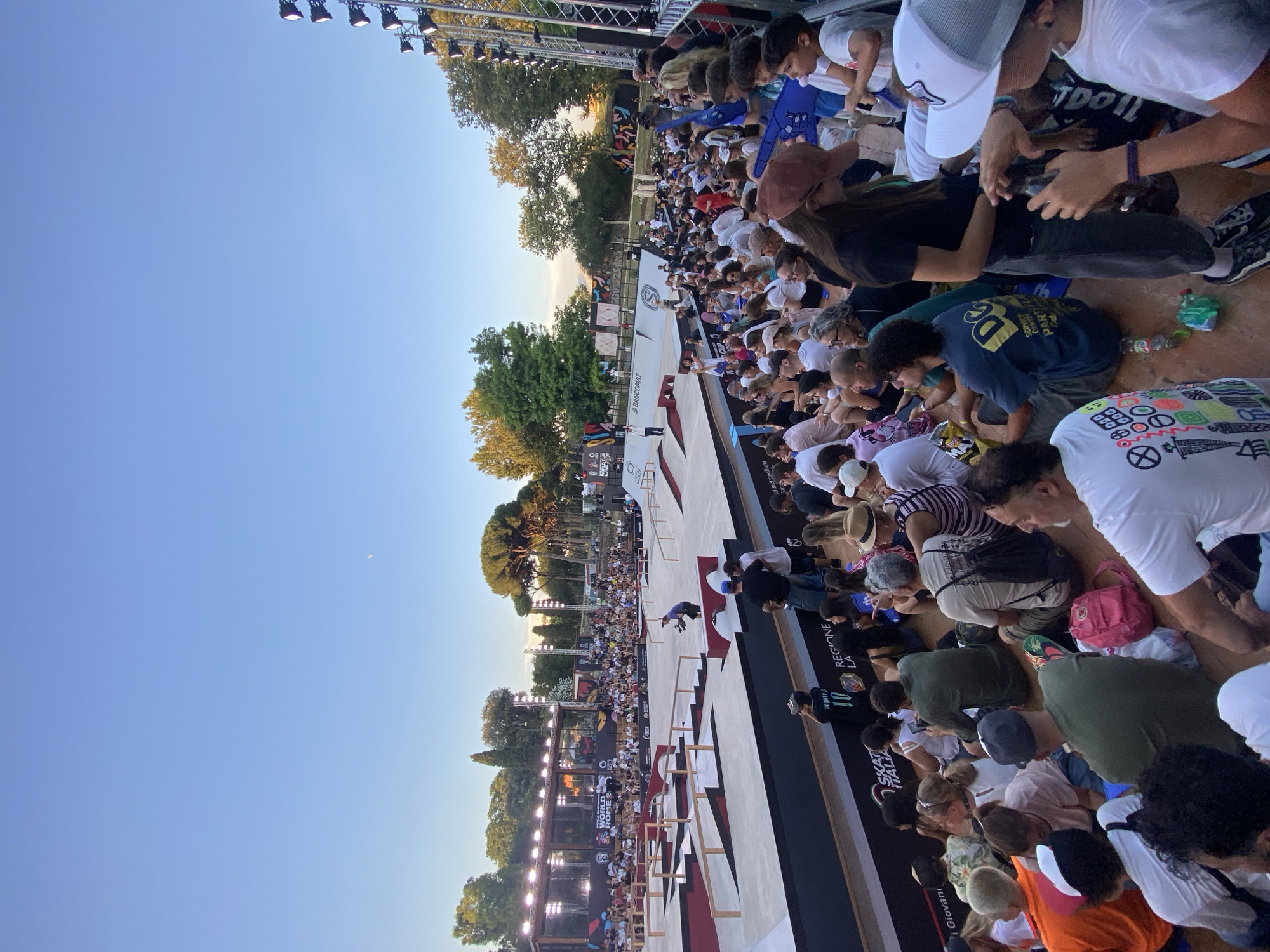 This image depicts a skateboarding event at the Skate World Rome, in Rome, Italy. The scene shows a large crowd of spectators seated on bleachers, watching a skateboarding competition on a complex course with ramps and obstacles. The event appears to be taking place outdoors during the daytime, with clear blue skies and sunlight illuminating the scene. The spectators are a diverse group of people of various ages, dressed in casual attire. Some are holding foam fingers and others are taking pictures with their phones. The skateboarding course is made of concrete and wood, with bright graphics and branding. The background features trees and buildings, suggesting an urban environment. The overall atmosphere seems to be one of excitement and engagement, with the crowd fully immersed in the event.