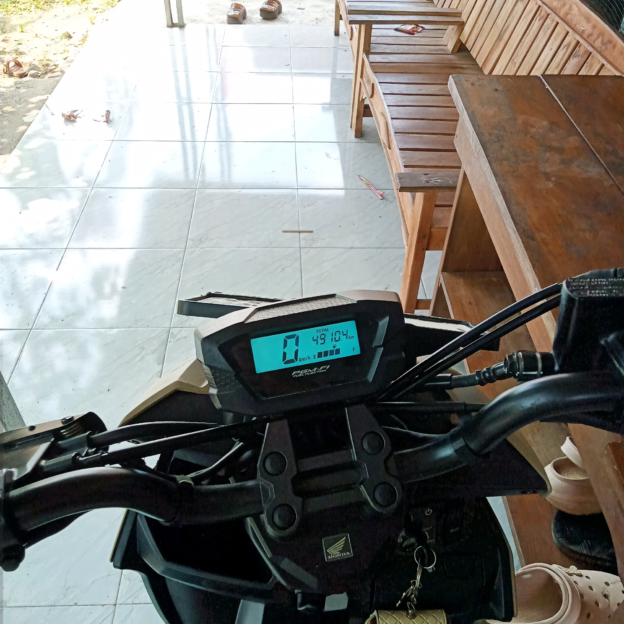 The image shows a close-up view of a motorcycle's digital dashboard displaying a speed of 0 km/h and a total distance of 49,104 km. The dashboard is on a Honda motorcycle, indicated by the "HONDA" logo visible below the display. The motorcycle is parked on a white tiled floor, possibly outdoors, as suggested by the presence of sunlight and shadows. In the background, a wooden table and chairs are visible, along with some scattered sandals on the floor. The scene appears to be in a residential or communal outdoor area, possibly in Purwakarta, Indonesia, given the context provided. There are no people directly visible in the frame.