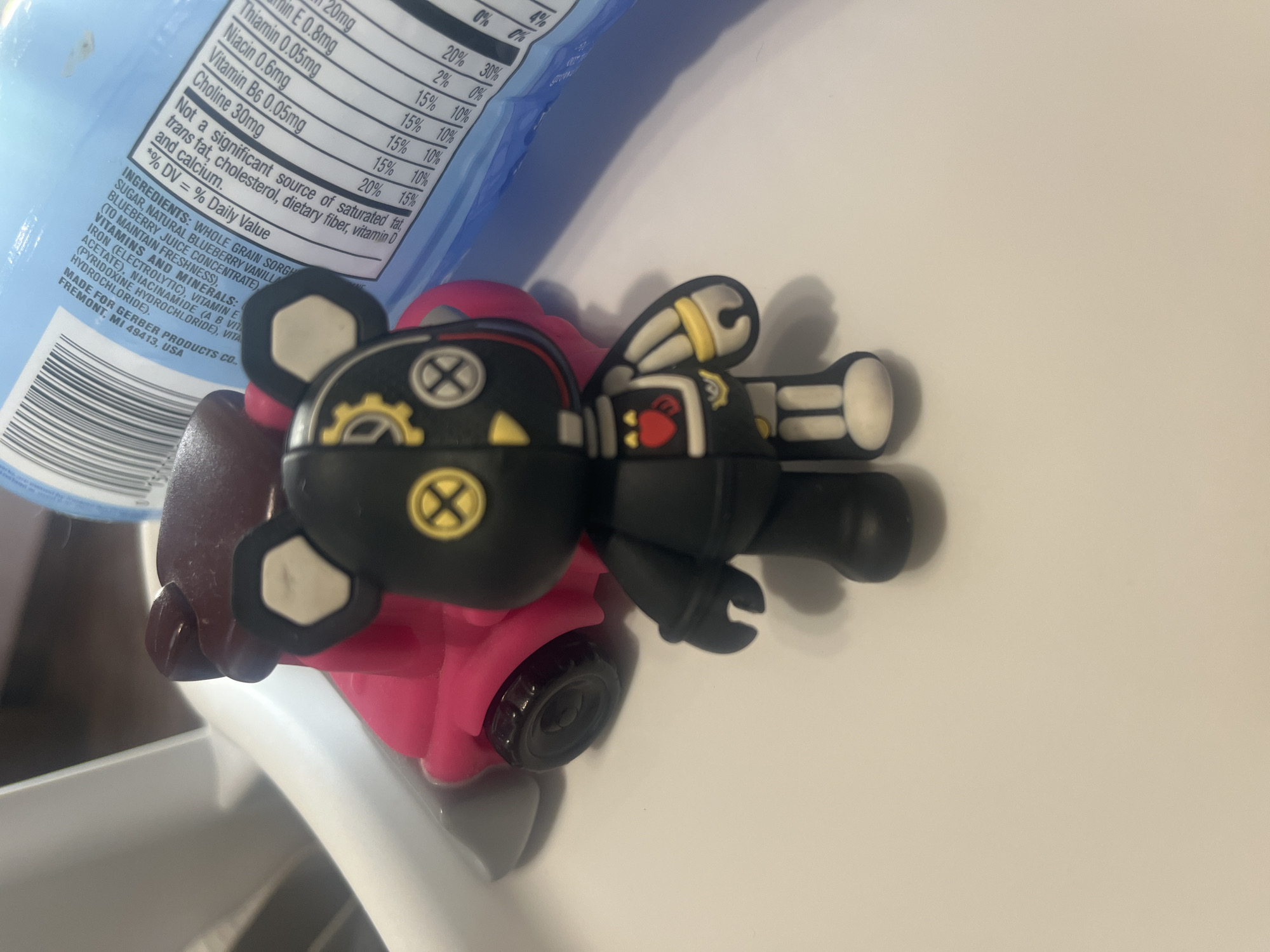 The image displays an indoor scene featuring a light blue flexible food pouch alongside a black, red, and yellow figurine, all resting on a white, flat surface. The pouch, likely for a food product, occupies the left side of the frame and displays nutritional facts and ingredients.
Visible text on the pouch includes nutritional values for Thiamin (0.05mg), Vitamin E (0.8mg), Niacin (0.6mg), Vitamin B6 (0.05mg), and Choline (30mg), along with daily value percentages. It explicitly states, "Not a significant source of saturated fat, trans fat, cholesterol, dietary fiber, vitamin D and calcium." The ingredients section mentions "WHOLE GRAIN SORGHUM," "SUGAR, NATURAL BLUEBERRY VANILLA," and "BLUEBERRY JUICE CONCENTRATE," among other vitamins and minerals like Iron, Vitamin E Acetate, Niacinamide, and Pyridoxine Hydrochloride. The origin is clearly marked as "MADE FOR GERBER PRODUCTS CO., FREMONT, MI 49413, USA." A partial barcode is also visible.
To the right, a black figurine, styled like a mechanical or steampunk bear or character, stands on a bright pink object that appears to be a small toy vehicle or base, complete with a black wheel. The figurine has light beige/white details on its ears, hands, and feet. It features yellow circular symbols (resembling an 'X') on its head and stomach, a red heart on its chest, and yellow/beige wing-like or gear-like structures on its back. The arrangement suggests an everyday, casual setting, possibly on a kitchen counter or table. No individuals are present in the image, and no specific time of day or external location cues are discernible.