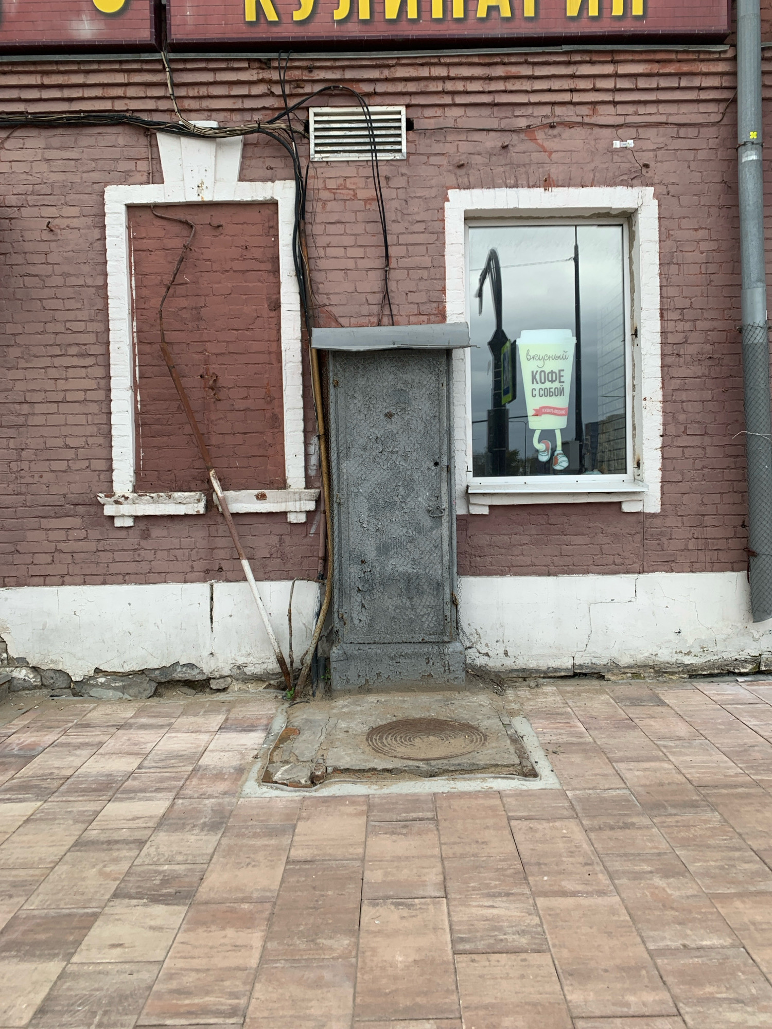 This image depicts the facade of a brick building located in Arkhangelsk, Russia. The building has a weathered appearance, with peeling paint on the white trim around the windows and door.
The left side of the building features a boarded-up window with a reddish-brown material, framed by white trim. A rusty metal rod leans against this window, extending from the ground to the top of the frame. Above this window, a white, raised architectural element is visible, with a white vent on the brick wall to its right.
In the center of the building is a gray, metal door, recessed slightly into the wall. The door has a rough, distressed texture, and appears to be old and worn. In front of the door, there is a metal manhole cover set into the pavement.
To the right of the door is a window with white trim. A sticker is visible on the glass, featuring a cartoonish depiction of a coffee cup with legs and shoes, and text in Russian that reads "вкусный КОФЕ С СОБОЙ" (delicious coffee to go). The window reflects the sky and some surrounding buildings.
The building's foundation is a rough, cracked concrete base, with some visible stones and dirt. The ground in front of the building is paved with rectangular, tan and brown interlocking pavers, laid in a running bond pattern.
The overall scene suggests an older urban environment, possibly a commercial building with some signs of neglect and age. The lighting indicates it is daytime, with overcast skies. There are no people visible in the image. The building's facade is cluttered with visible electrical wiring running along the brickwork.