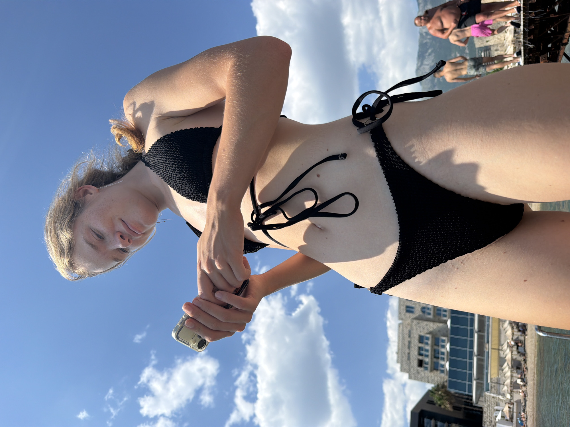 This is a low-angle, portrait-oriented, outdoor photo taken on a sunny day, likely in the afternoon, given the lighting and shadows. The primary subject is a young woman in a black bikini, seen from the waist up, with her head tilted back and looking downwards towards the camera. Her blonde hair is pulled back, and she is holding a smartphone in her hands, which are positioned in front of her. The background shows a clear blue sky with scattered white clouds, and a large, modern building with many windows and balconies is visible further in the distance, along with beach umbrellas and people. There is also a hint of water and mountains in the far background, suggesting a coastal location. The overall mood is relaxed and casual, consistent with a day at the beach or by the water. No visible text is present in the image. The scene appears to be set in Budva, Montenegro, a popular tourist destination known for its beaches and scenery.