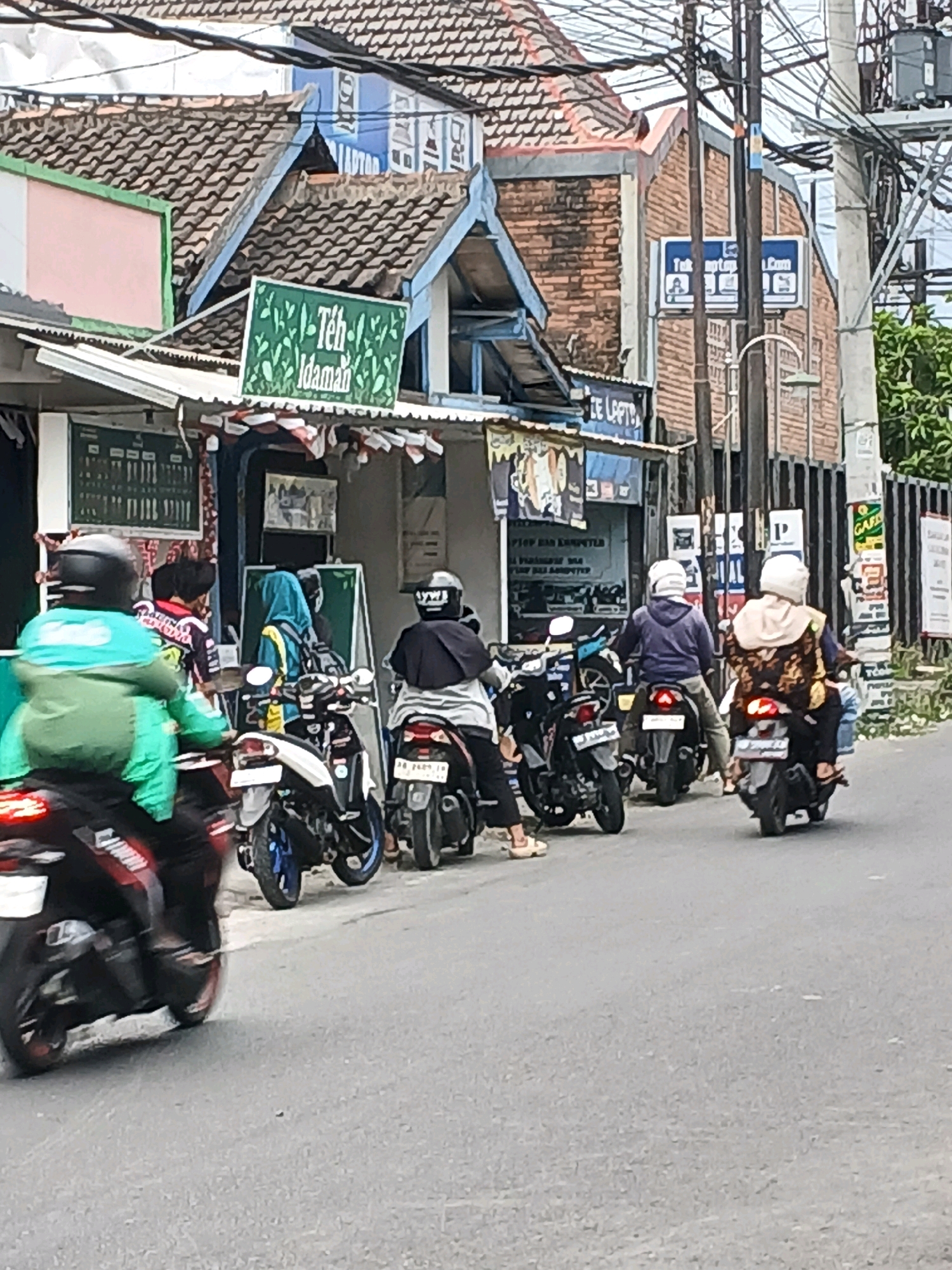 In Depok, Indonesia, a street scene unfolds with several individuals on motorcycles, indicating a common mode of transportation in the city. The foreground is dominated by the rear of a motorcycle with a rider wearing a bright green jacket and a black helmet, suggesting they are either in motion or paused. Behind them, a line of other motorcycles and riders can be observed, some with passengers. The urban environment is characterized by buildings with tiled roofs, overhead wires, and street poles, typical of Indonesian streetscapes. A sign in green with white lettering, reading "Teh Idaman," is visible on one of the buildings, likely indicating a tea shop or beverage stall. The overall impression is of a busy street, possibly during the day, with daily life and commerce taking place. The presence of multiple motorcycles suggests traffic or a gathering point. The buildings appear to be a mix of residential and commercial structures, common in the bustling urban areas of Depok.