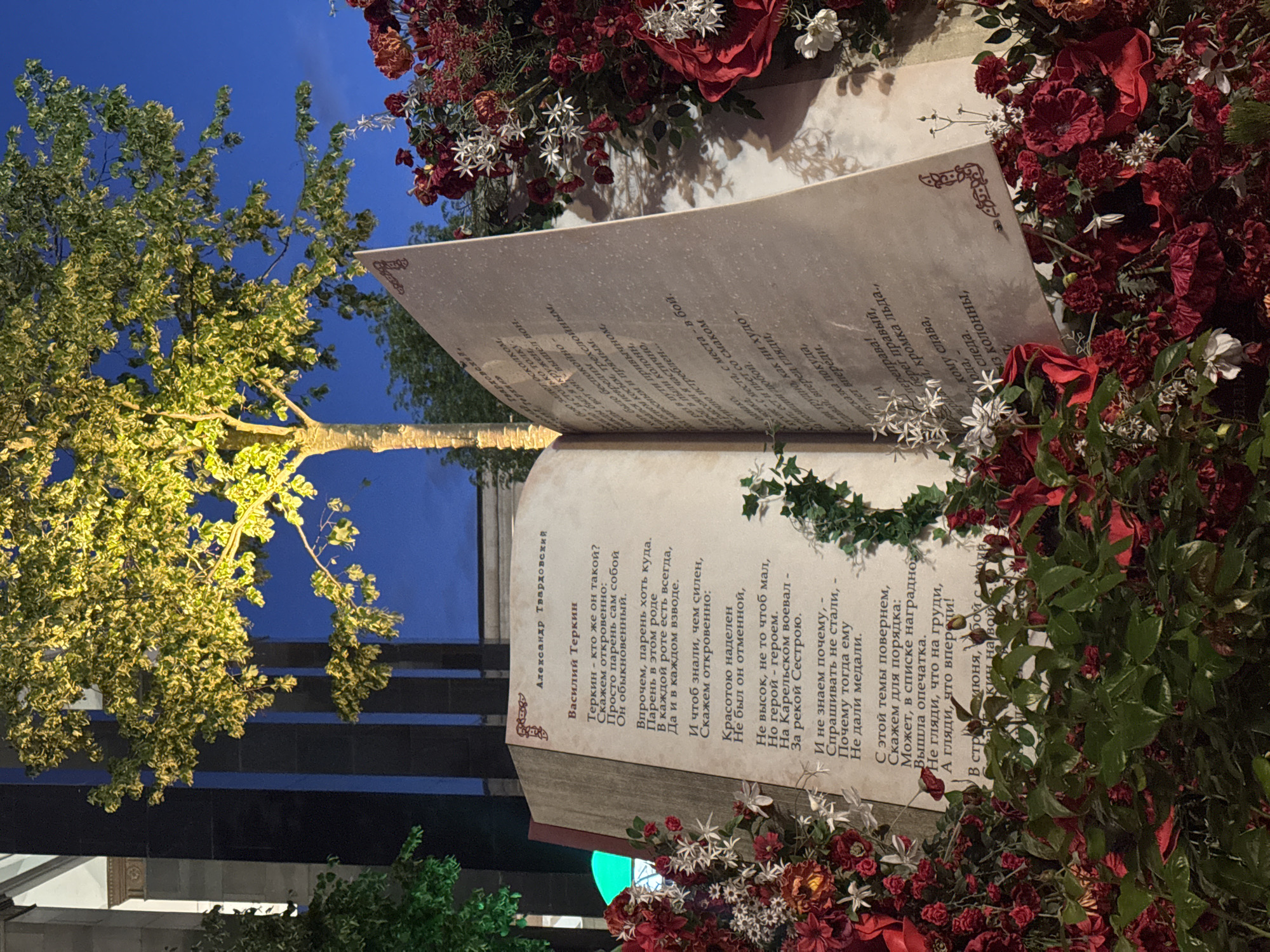 The image shows an open book with text on its pages, placed outdoors amidst a floral arrangement of red and white flowers and greenery. The book appears to be made of stone or a similar material and is designed to resemble a large, open volume. The text on the left page is in Russian and appears to be a poem. Above the text on the left page, the name "Александр Твардовский" (Alexander Tvardovsky) is visible. Below that, the title "Василий Теркин" (Vasily Terkin) is present. The text on the right page is also in Russian and seems to be a continuation of the poem.
The background shows a dark, modern building with vertical columns, possibly made of dark stone or metal. A large tree with illuminated yellow-green leaves is positioned behind and above the book, creating a dramatic lighting effect. The sky in the background is a deep blue. The overall scene suggests an outdoor exhibition or art installation.
Based on the context of Alexander Tvardovsky's "Vasily Terkin," a famous Soviet poem about a soldier in World War II, and the style of the installation, this scene is likely from an exhibition related to literature, history, or a commemorative event. Without additional information, pinpointing the exact location in Moscow is difficult, but it is plausible to be a public space like a park, square, or a cultural institution's grounds.