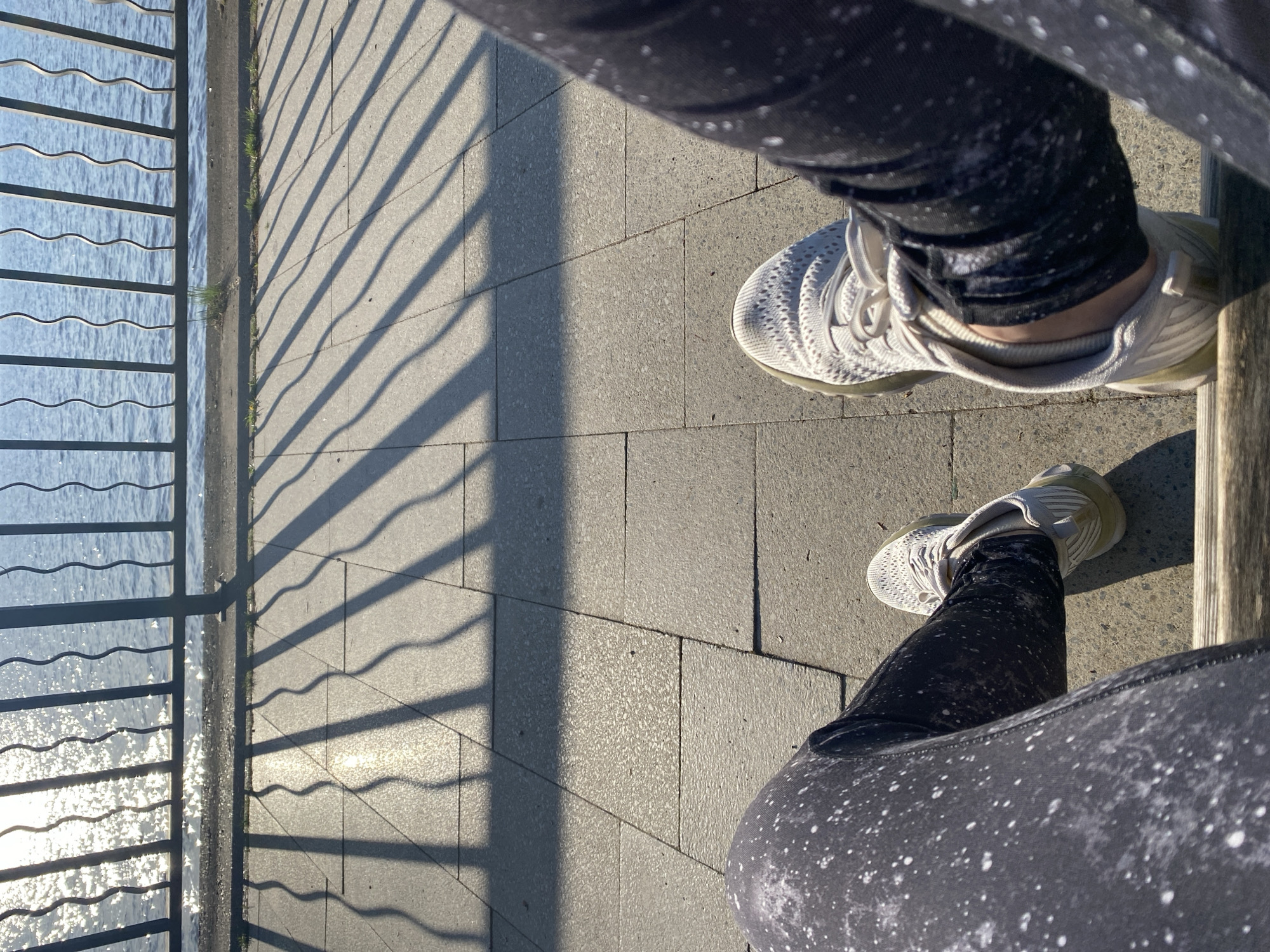 The image shows a first-person perspective of someone standing on a paved area next to a body of water, likely a river or lake, bordered by a metal fence. The water reflects sunlight. The person is wearing black leggings with white speckles and off-white sneakers. Shadows from the fence are cast onto the pavement and leggings. The location is likely Chaikovsky, Russia. There's a wooden beam beside the person's right foot, suggesting they are sitting on a bench.