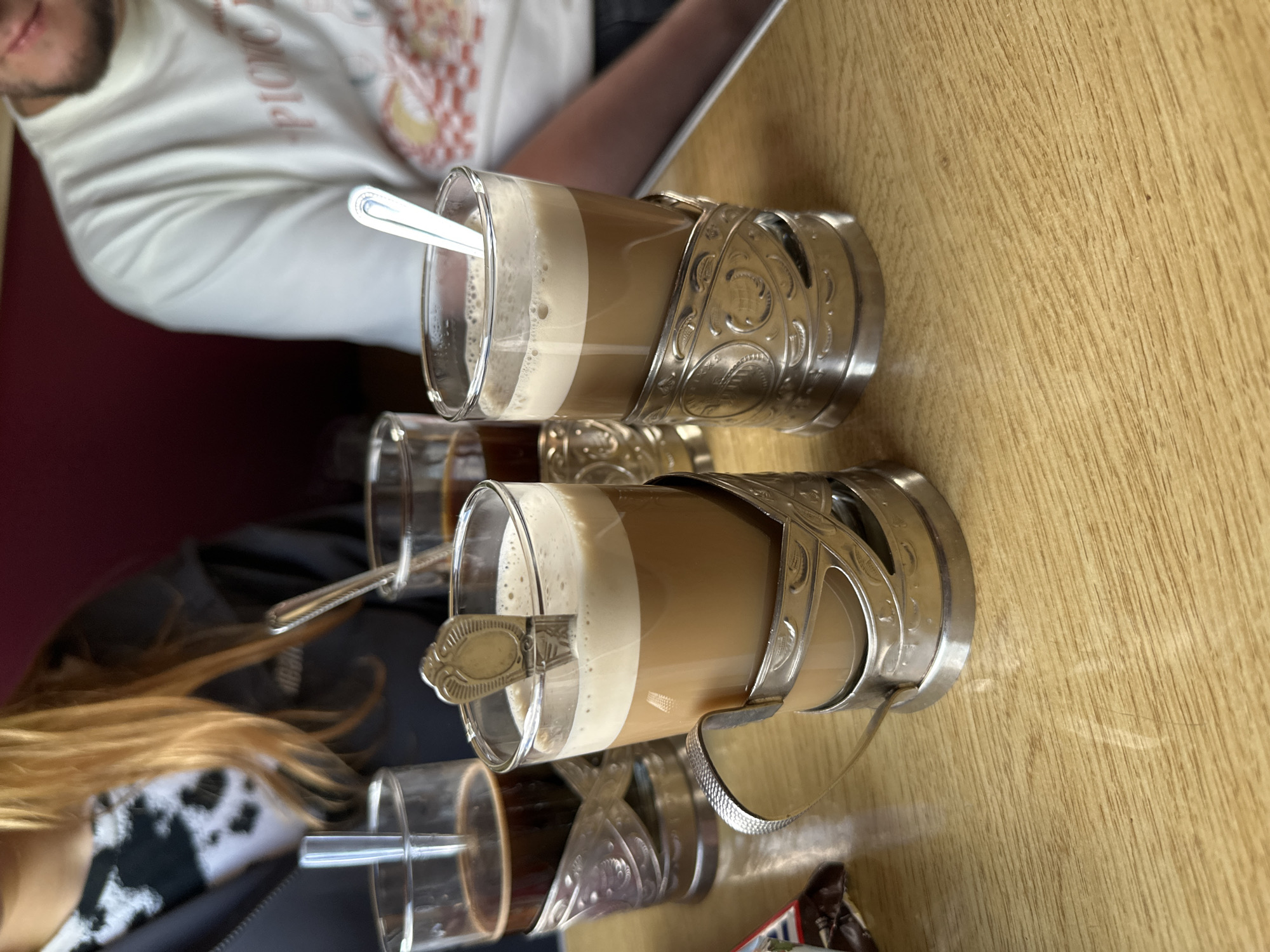 The image shows two glasses of layered coffee drinks served in ornate silver-colored metal holders on a wooden table. Each glass has a spoon inside. Two additional, smaller glasses containing dark liquids are also visible in the background. A person wearing a light-colored patterned shirt is partially visible in the upper left, and another person wearing a black and white patterned garment is partially visible in the lower left. The photo was taken indoors. While the location is indicated as Мажэйкаўскі сельскі Савет, Belarus, there are no visual cues in the image to confirm this specific location within the city.