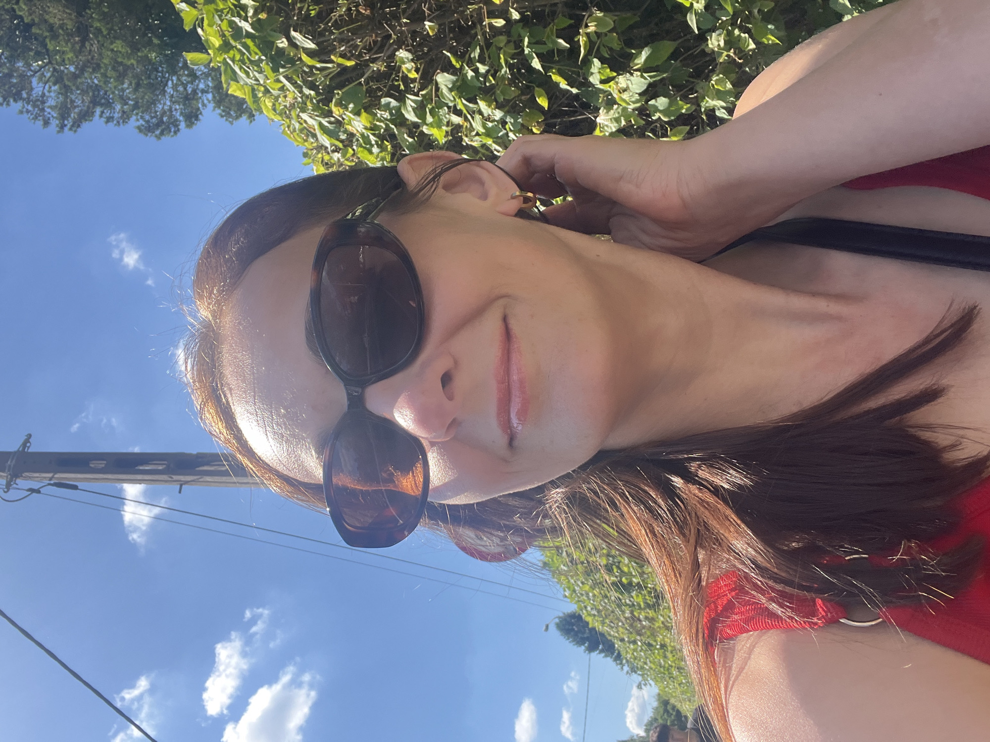 A woman with brown hair and sunglasses is taking a selfie outdoors on a sunny day. She is wearing a red top and a gold hoop earring. She is holding her hand to her ear and smiling. Behind her, there is a wooden utility pole, lush green bushes, and a blue sky with white clouds. The location is not specified.