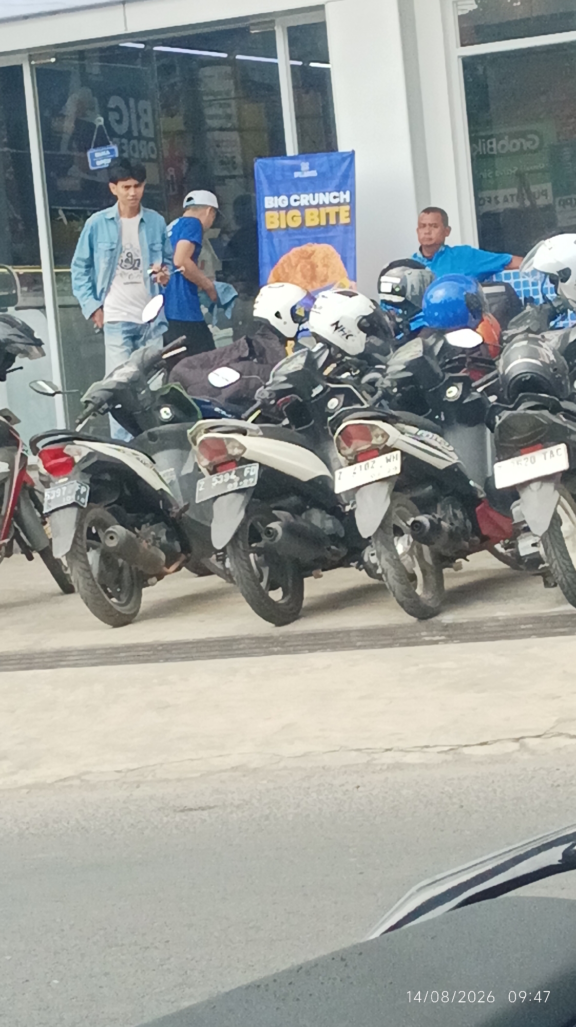 The image depicts an outdoor scene in Tasikmalaya, Indonesia, possibly in front of a store or commercial establishment, as indicated by the signage "BIG CRUNCH BIG BITE." The foreground features a row of parked motorcycles, mostly scooters, with several helmets placed on them. The motorcycles appear to be of common Indonesian brands.
In the background, three men are visible. Two men stand near the entrance of a building. One man, wearing a light blue denim jacket over a white t-shirt and blue jeans, is looking towards the viewer. Another man, in a blue t-shirt and a white cap, is holding a cloth or rag in his right hand, and his gaze is directed downwards, possibly cleaning something. Further to the right, a third man, wearing a blue shirt, is leaning against a white surface, looking towards the left side of the frame.
The environment suggests a commercial area, with a building facade and windows visible. The ground is paved, and the overall lighting indicates it is daytime. The date and time stamp "14/08/2026 09:47" is present in the lower right corner, but this may be a timestamp from the camera and not indicative of the actual date of the photograph. The presence of multiple motorcycles suggests a location that might attract customers who arrive by vehicle.