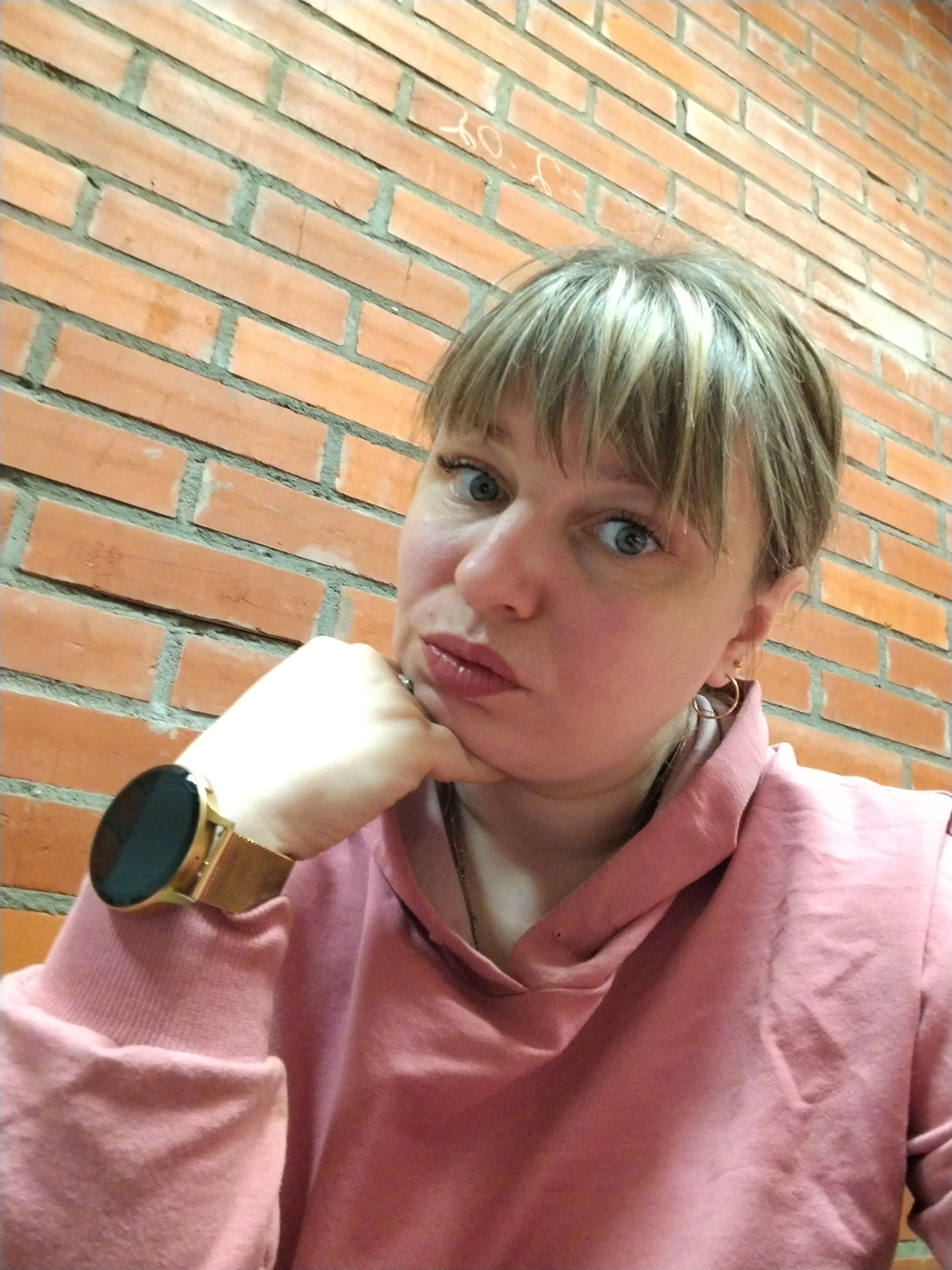 A close-up selfie depicts an adult woman with light blonde hair and bangs, and light blue-green eyes. She is wearing a pink or salmon-colored hooded sweatshirt. Her left hand, adorned with a gold-colored smartwatch featuring a black screen and a mesh band, rests against her chin with her fingers lightly curled. A small gold hoop earring is visible in her right ear. Her lips are pursed, and she looks directly at the camera with a thoughtful or slightly pouting expression. The background is a textured orange-red brick wall with grey mortar. Faint, illegible light-colored markings or scribbles are visible on some of the bricks in the upper left portion of the image. The setting is in Шебунино, Russia, likely indoors or against an outdoor brick facade, with no specific time of day or weather discernible from the lighting.