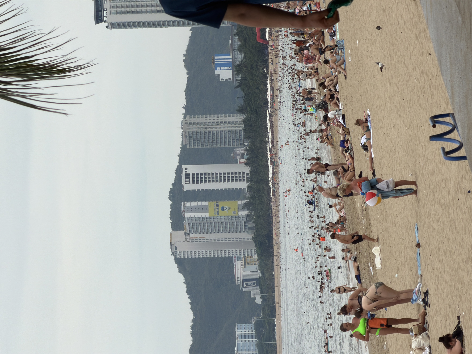 This is a wide shot of Nha Trang beach in Vietnam, bustling with activity. The foreground is dominated by a sandy beach populated with numerous people engaged in various activities. Several individuals are sunbathing on towels, while others are walking or standing. Closer to the water, groups of people are wading and swimming in the ocean. Numerous umbrellas are set up along the beach, providing shade for beachgoers. In the middle ground, the ocean is filled with a large number of people, some swimming, others on inflatable devices or small boats. The background reveals a dense line of palm trees separating the beach from a series of tall, modern buildings. Several high-rise hotels and apartments are visible, some with balconies and windows. The buildings are situated against a backdrop of lush green hills, indicating a coastal urban environment. The sky is overcast and a light gray, suggesting cloudy weather. In the far distance, a few more buildings and the hazy outline of mountains can be seen. The scene depicts a lively beach resort atmosphere, characteristic of popular tourist destinations in Nha Trang.