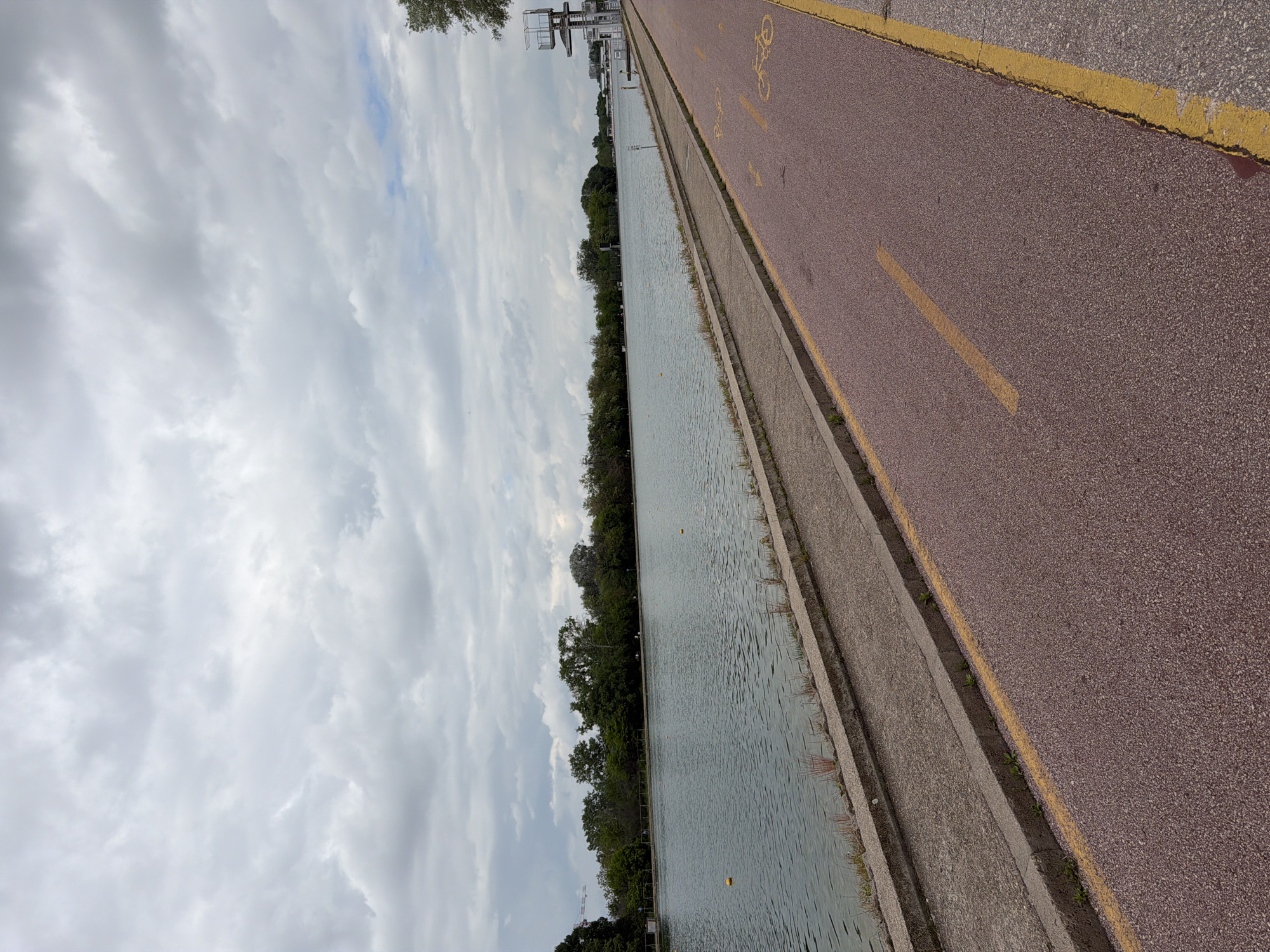 The image shows a long, narrow body of water next to a paved path. The path, designed for cyclists, has a reddish hue and yellow markings. A concrete barrier separates the water from the path. Along the water's edge, there's a line of trees. The sky is overcast with heavy clouds. The scene depicts the rowing channel in Plovdiv, Bulgaria.