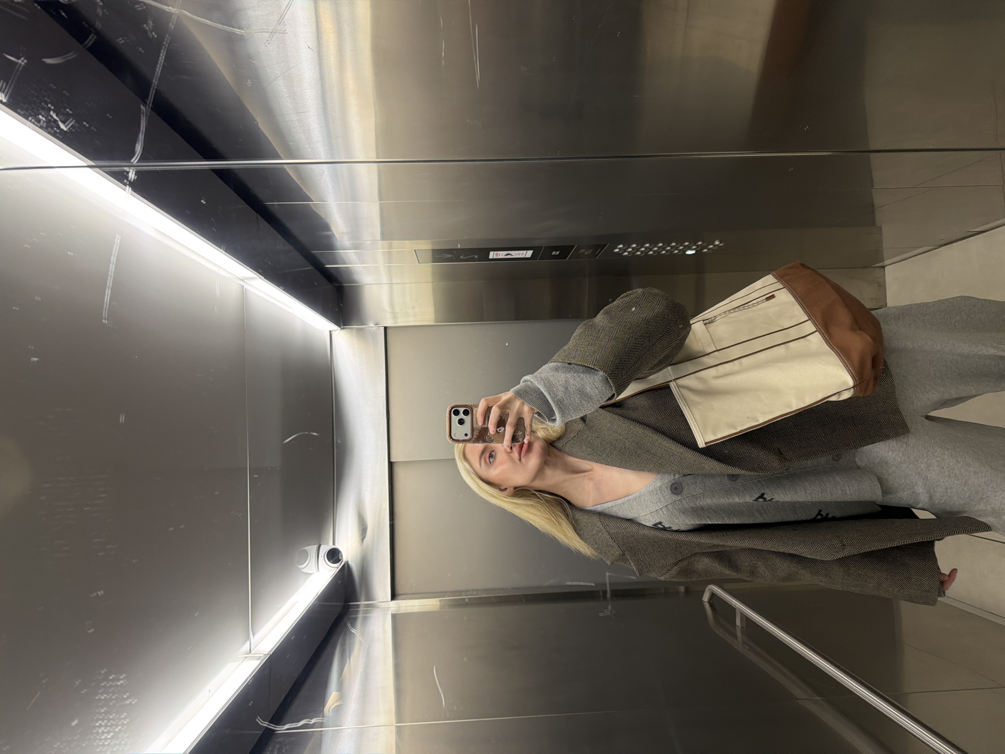 A young blonde woman is depicted taking a selfie in a reflective elevator in Belgrade, Serbia. The image is captured through the reflection of a mirror or highly polished metallic wall, with the woman holding her phone up to capture her reflection. She is wearing a gray long-sleeved top, partially obscured by a dark brown or olive green blazer with a herringbone pattern. A cream-colored tote bag with brown accents hangs from her shoulder. Her expression is neutral, with a slight pout, as she looks directly at her phone. Her long blonde hair falls over her shoulders.
The elevator interior features sleek, metallic walls, likely brushed stainless steel, which highly reflect the interior lights and the woman. Horizontal strip lighting illuminates the space. In the upper right corner of the image, a small security camera is visible. The elevator control panel is visible in reflections and directly above the woman's head, displaying various buttons and symbols, including door open/close indicators and an 'S' symbol, though specific numbers are indistinct. The surfaces show some scratches, indicating wear. The woman's lower body is clad in gray trousers, possibly sweatpants, with a small portion of her feet visible, appearing to be bare or in light-colored footwear. The overall setting suggests a modern, functional environment.