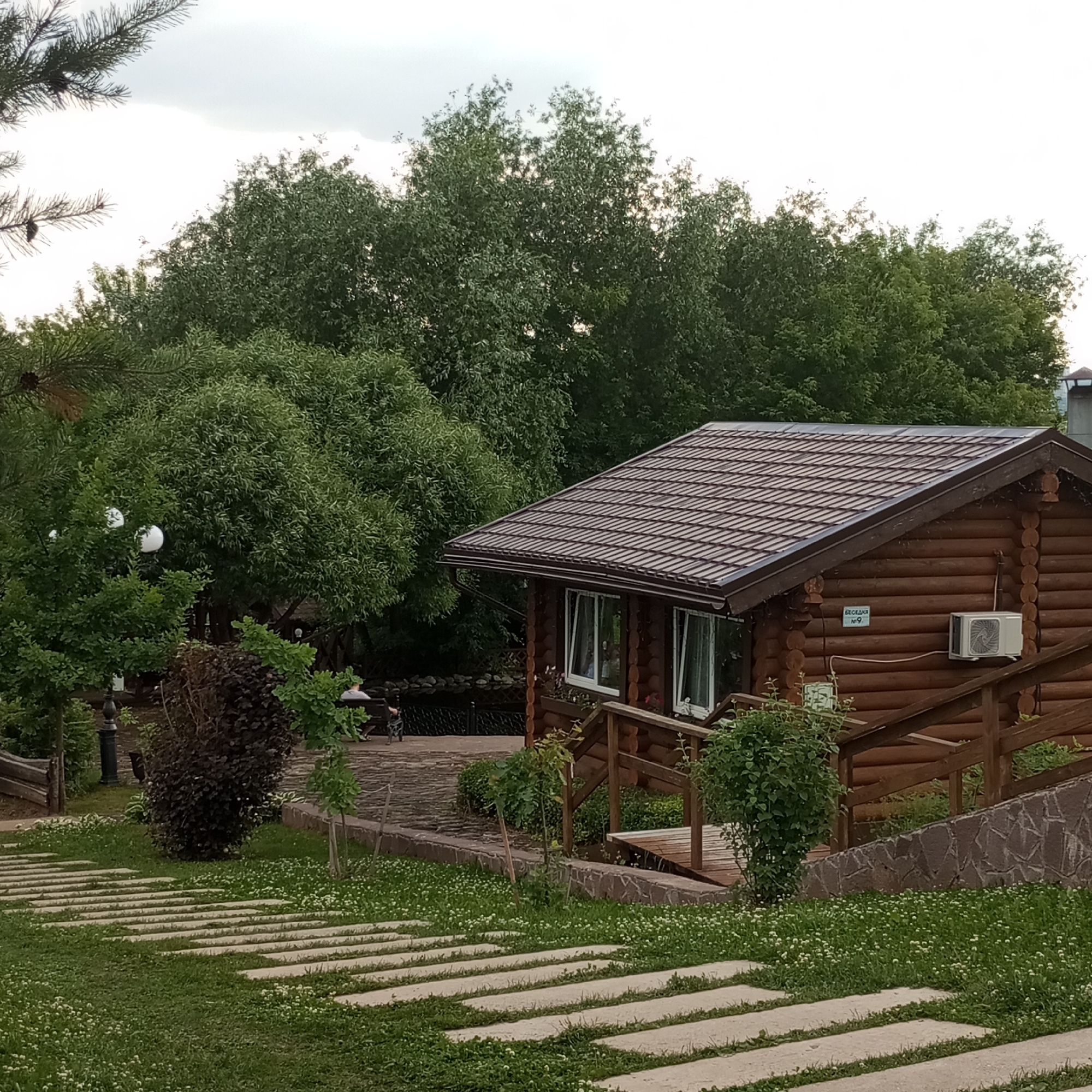 The image depicts a rustic wooden cabin with a brown metal roof, set amidst lush greenery and trees. A stone pathway leads to the cabin, which has two white-framed windows and an air conditioning unit mounted on its exterior wall. A sign with Russian text, "Беседка №9" (Gazebo No. 9), is visible on the cabin's wall. A wooden staircase with railings provides access to the cabin's entrance. The surrounding area features manicured lawns with small white flowers, bushes, and a paved area with benches. A body of water, possibly a pond or lake, is visible in the background, with a fence separating it from the cabin area. The sky is overcast. In the foreground, there is a pathway made of concrete slabs interspersed with grass. The scene appears to be a tranquil park or recreational area.