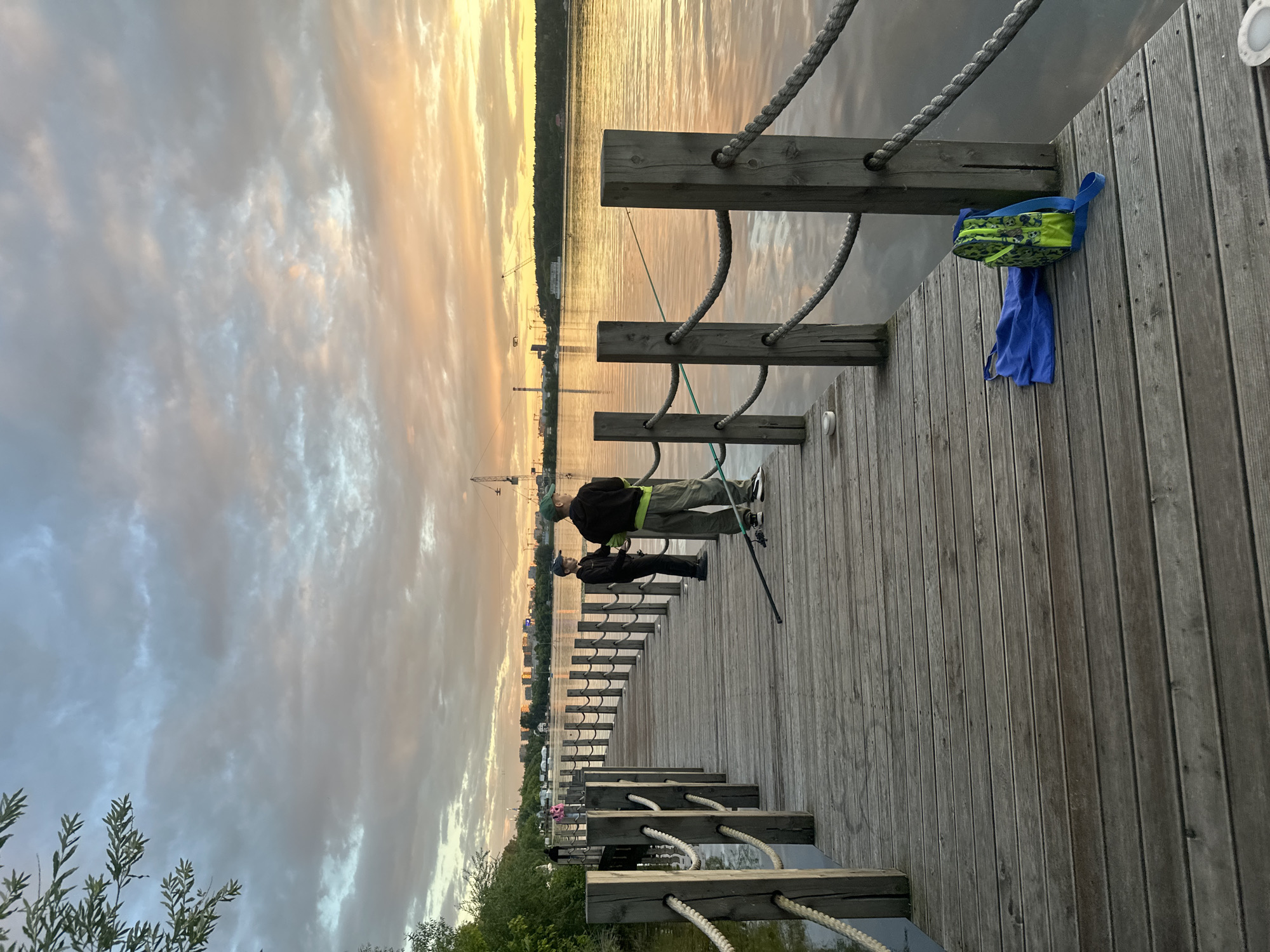 The image shows two people fishing from a wooden pier at sunset. The sky is filled with dramatic clouds painted in shades of orange, yellow, and grey, reflecting on the calm water. In the background, a city skyline with cranes suggests an urban or industrial area. The pier is made of weathered wooden planks and has rope railings. On the pier, a green and blue bag and a blue towel hang from a wooden post. The overall atmosphere is serene, with the warm light of the setting sun creating a peaceful mood. The scene appears to be in Myakinino, Russia, as indicated by the provided location context.