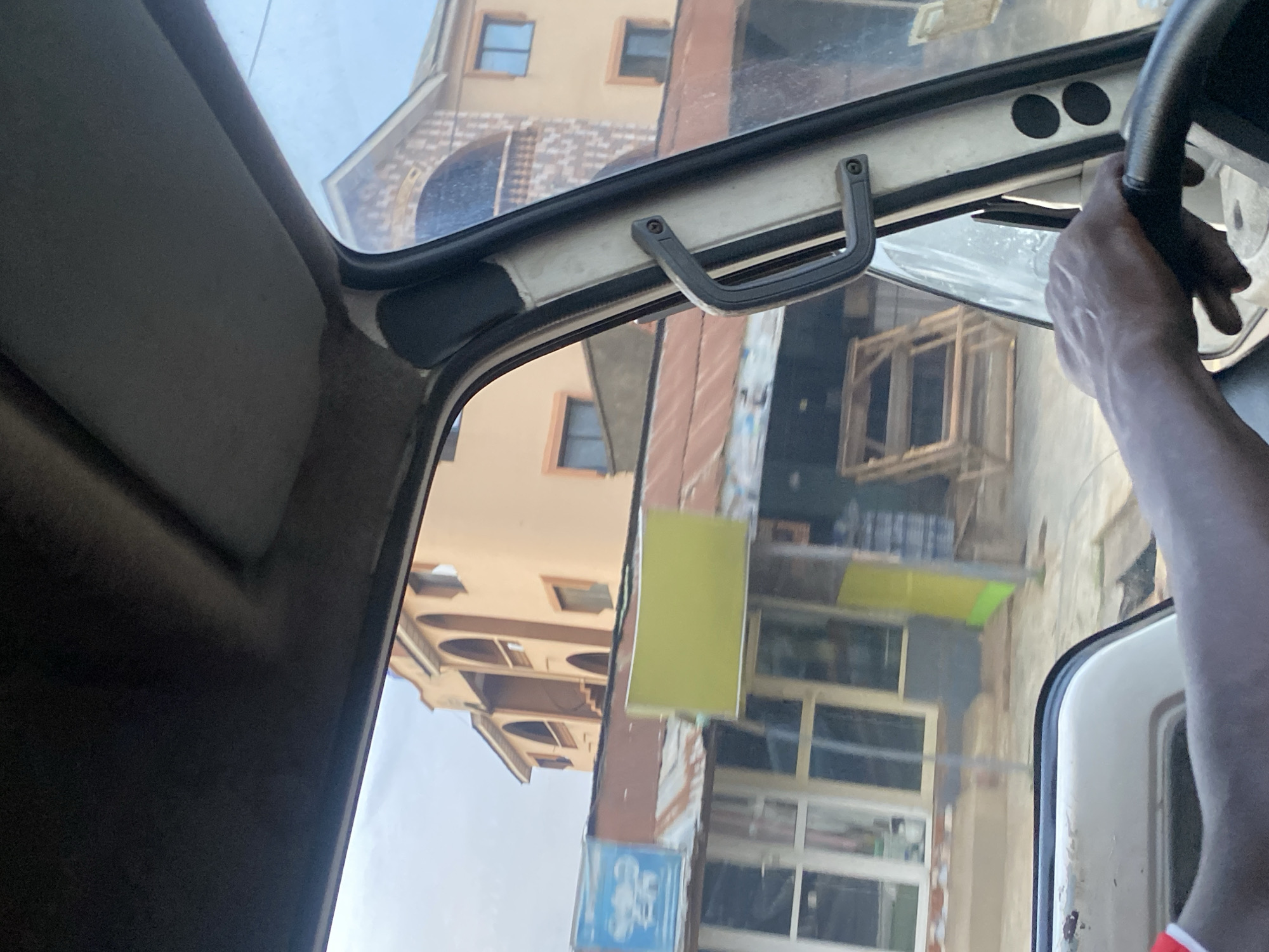 The image provides an interior view from the driver's seat of a vehicle, looking out through the windshield and side window. The perspective suggests the vehicle is in motion, or has recently stopped, on a street in Ikorodu, Nigeria. In the foreground, a dark-skinned hand is visible on the steering wheel, indicating the presence of a driver. The interior of the vehicle is sparsely detailed, but the dashboard and part of the roof lining are visible.
Outside the vehicle, the environment is characteristic of a busy urban street in Nigeria. Directly ahead and to the left, a two-story building with arched windows and balconies is prominently featured. The building's facade is painted in a light peach color. In front of this building, there are several shops with corrugated metal roofs, some of which appear weathered. One shop has a blue sign with white lettering and graphics, and another has a large, unlettered, bright yellow rectangular sign. The street itself is paved, and there are indications of foot traffic and other vehicles further down. To the right, stacked wooden frames or pallets are visible outside one of the establishments, suggesting commercial activity. The sky appears overcast, typical of many days in the region. The overall scene captures a slice of everyday life in a commercial district of Ikorodu.