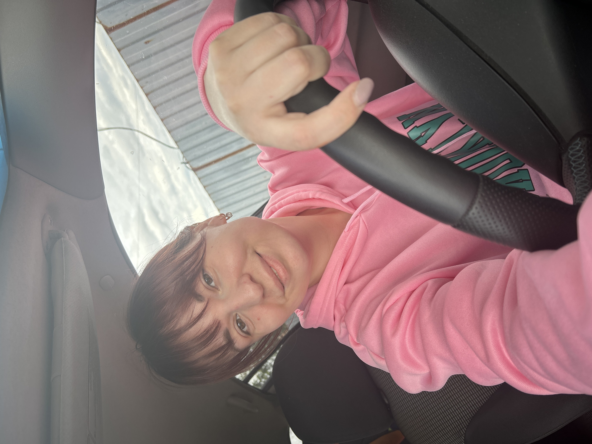 A woman with brown hair and bangs, wearing a pink hoodie, is holding a black steering wheel inside a car. The car is parked. The background shows a blurry outdoor scene, including what appears to be a corrugated metal roof or structure. The location is likely in the city of Фоки, Russia.