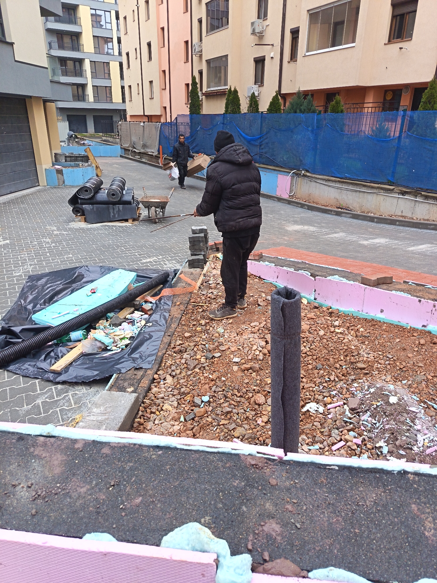 The image captures an outdoor construction site in what appears to be Sofia, Bulgaria, given the architectural style of the surrounding buildings. Two workers are present. One, further away, carries a cardboard box. The other, closer to the foreground, is placing brick pavers. Construction materials are visible, including rolls of black roofing material, a wheelbarrow, insulation boards (pink and blue), a large black plastic sheet covering construction debris, drainage pipes, and gravel. The ground is uneven and shows signs of ongoing work. A blue construction fence is also visible in the background.