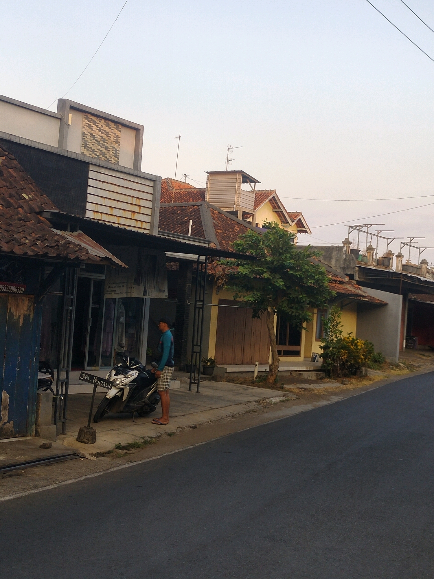A man stands next to a motorcycle on the sidewalk in front of a building in Pujut, Indonesia. The man is wearing a blue long-sleeved shirt and checkered shorts. The motorcycle is white and black with a license plate that reads "3985 JBK - 12 - 23". There is a sign in front of the motorcycle that says "JUAL PERTALIL". The building behind the man appears to be a shop with a glass door and windows. There is a tree in front of the shop, and other buildings can be seen in the background. The sky is clear and a light blue.