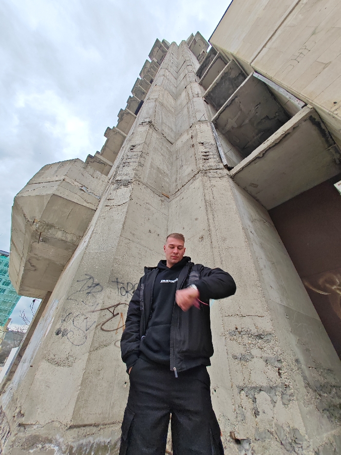 An outdoor, low-angle photograph depicts a male individual standing directly in front of the unfinished Hotel 'Shumen', a brutalist concrete building located in Shumen, Bulgaria. The subject, a male with short, light-colored hair and a light complexion, is dressed in a black hooded sweatshirt featuring a white graphic, a black puffer jacket, and black cargo pants. He stands with his left hand in his pocket and is looking downwards at an object on his left wrist, which appears to be a watch. The large, multi-story concrete structure dominates the background, characterized by its exposed, raw concrete facade and numerous rectangular, cantilevered sections. The lower walls of the building display black spray-painted graffiti. The sky is overcast. To the far left in the background, another structure partially obscured by light blue or green construction netting is visible.