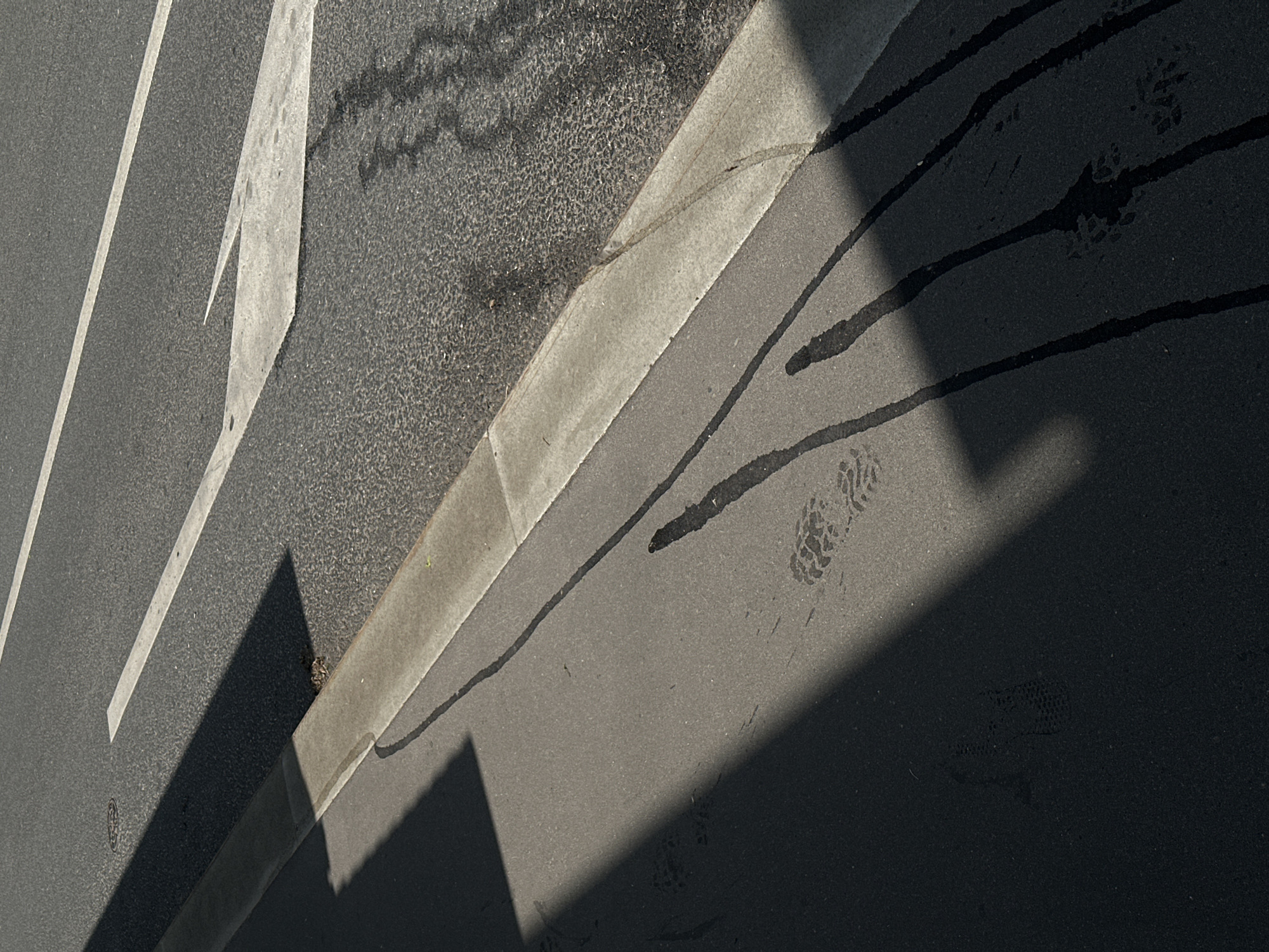 The image shows an urban street scene in Moscow, Russia. The ground is asphalt, marked with white painted lines and directional arrows. A concrete curb separates the street from a different textured asphalt surface. Shadows from a building or object fall diagonally across the scene, creating a strong contrast. Dark stains or drips are visible on the street, appearing to originate from the direction of the shadow. A partial footprint can also be discerned on the street.