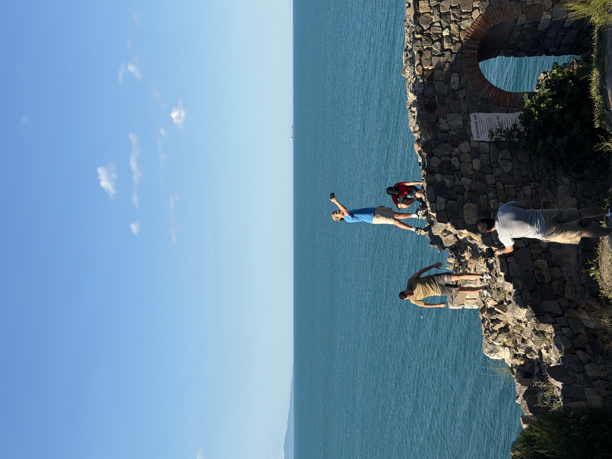 Four men are present on the ruins of an ancient structure overlooking the Black Sea in Sozopol, Bulgaria. The man on the far left is wearing a tan t-shirt and shorts, with a baseball cap. He is looking away from the camera and appears to be walking. The second man from the left is wearing a blue t-shirt and shorts. He is holding a smartphone and taking a picture, with his arm extended. He is standing on the edge of the ruins. To his right, a man in a red shirt and dark shorts is crouching and looking down. The fourth man, on the far right, is wearing a gray t-shirt and shorts. He is holding a smartphone and appears to be recording or taking a photo of the man in the blue shirt. The ruins are made of stone, with an arched opening on the right side that looks out towards the sea. A sign with text is mounted on the wall to the left of the arch. The sea is a deep blue, and a ship is visible on the horizon. The sky is clear blue with a few white clouds. The scene is lit by sunlight, suggesting it is daytime.