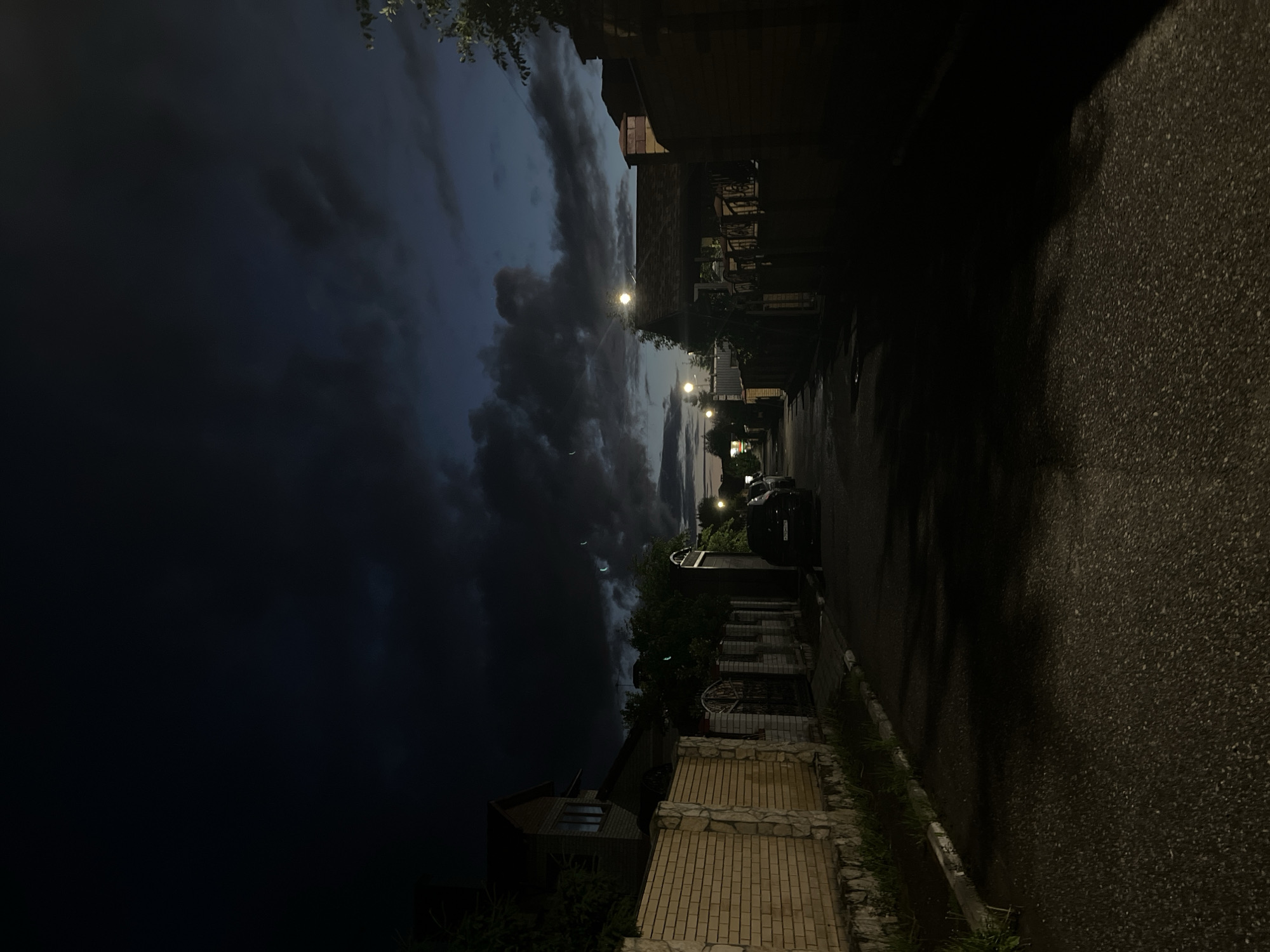 The image captures a residential street or alleyway in Ореховка, Russia, at night or late evening. The perspective looks down the street, with buildings on the left and a paved road on the right.
The sky, visible on the left, is filled with dramatic, dark, and heavy clouds, suggesting an overcast night or an impending storm. Several distinct crescent-shaped reflections, likely lens flares from light sources, are scattered across the dark cloud cover.
On the left side of the frame, a row of houses and buildings lines the street. One house in the foreground features a brick facade and a set of brick and stone steps leading up to it. Fences and some overgrown vegetation are also visible along the properties. Scattered trees and bushes dot the scene.
On the right, a dark, asphalt road stretches into the distance, appearing wet or reflective, possibly from recent rainfall. Several streetlights are illuminated, casting pools of light and creating stark shadows on the road and buildings. A dark-colored car is parked on the right side of the street further down.
The scene is devoid of human activity, conveying a sense of quietude or solitude. No visible text is legible on any of the objects, vehicles, or buildings. The overall atmosphere is dark and moody due to the low light conditions and heavy clouds.