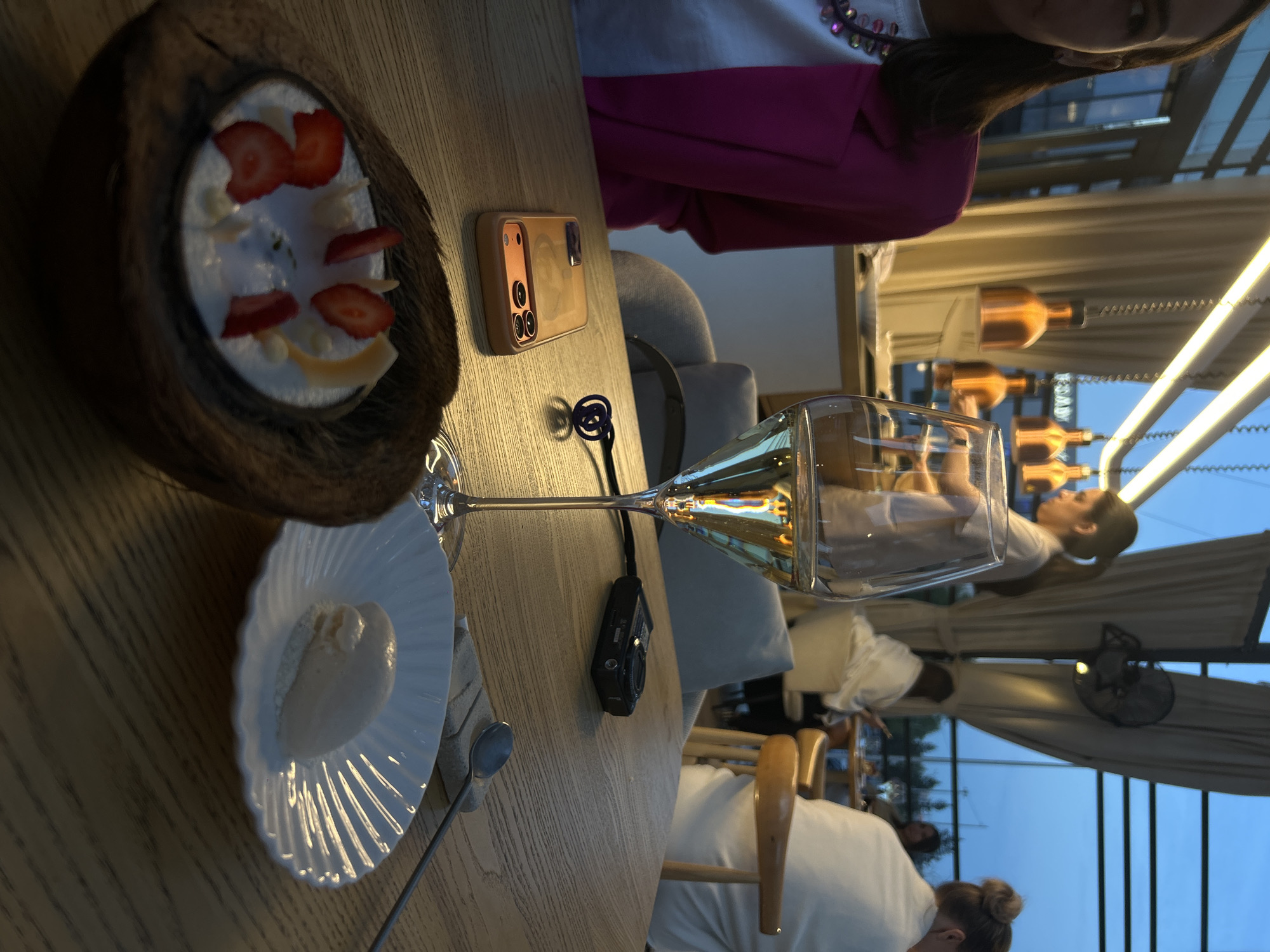 This image captures a vibrant dining scene in what appears to be a stylish restaurant or cafe, likely during late afternoon or early evening given the blue sky visible outside and the use of warm artificial lighting. The setting suggests an enclosed outdoor area, possibly a terrace or rooftop, with large glass panels or windows and light-colored curtains.
In the foreground, a wooden table is set with several items. On the left, a visually appealing dessert is presented in a natural coconut shell, filled with white cream, fresh sliced strawberries, and what appear to be flakes of white chocolate or dried fruit. Next to it, a white, fluted plate holds a scoop of a light-colored dessert, possibly ice cream or sorbet, accompanied by a spoon.
Towards the center of the table, a smartphone with a prominent camera array, enclosed in a light-colored case, rests face down. A small, black compact camera is also visible nearby. A tall, elegant wine glass, filled with a pale yellow beverage (likely white wine or champagne), stands prominently.
In the mid-ground and background, the restaurant's ambiance is evident. Warm, bottle-shaped pendant lights hang from the ceiling, casting a glow. Several people are present, indicating a busy dining environment. One person in the foreground, whose face is partially visible, wears a vibrant magenta jacket over a white top with colorful embellishments. Further back, a woman with dark hair tied back is seated at another table. Other patrons are visible in the soft focus of the background, contributing to the lively atmosphere.
The overall scene conveys a sense of leisurely enjoyment, with patrons partaking in desserts and drinks in a modern and inviting space. No specific text is clearly visible in the image.