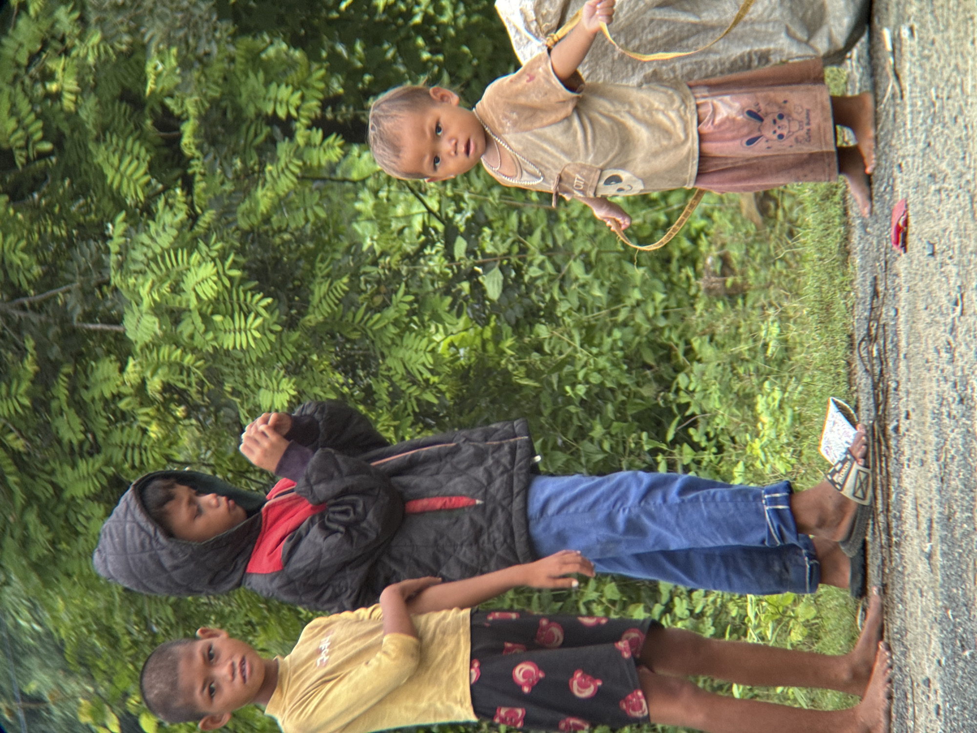 The image shows three children standing on a gravel road with lush green foliage in the background. The child on the left, a boy, is wearing a yellow long-sleeved shirt and black shorts with a pink patterned design. The middle child, also a boy, is wearing a black hooded jacket with a red lining and blue pants. He is looking down at his hands, which are clasped together. The child on the right, a younger boy, is wearing a light brown t-shirt with a cartoon print and light pink shorts. He has a necklace and is holding a yellow string or ribbon in both hands. Next to him is a large, light-colored bag. The setting appears to be outdoors, possibly on the outskirts of a rural area or a less developed urban part of Lao Bảo Commune, Vietnam. The lighting suggests it is daytime.